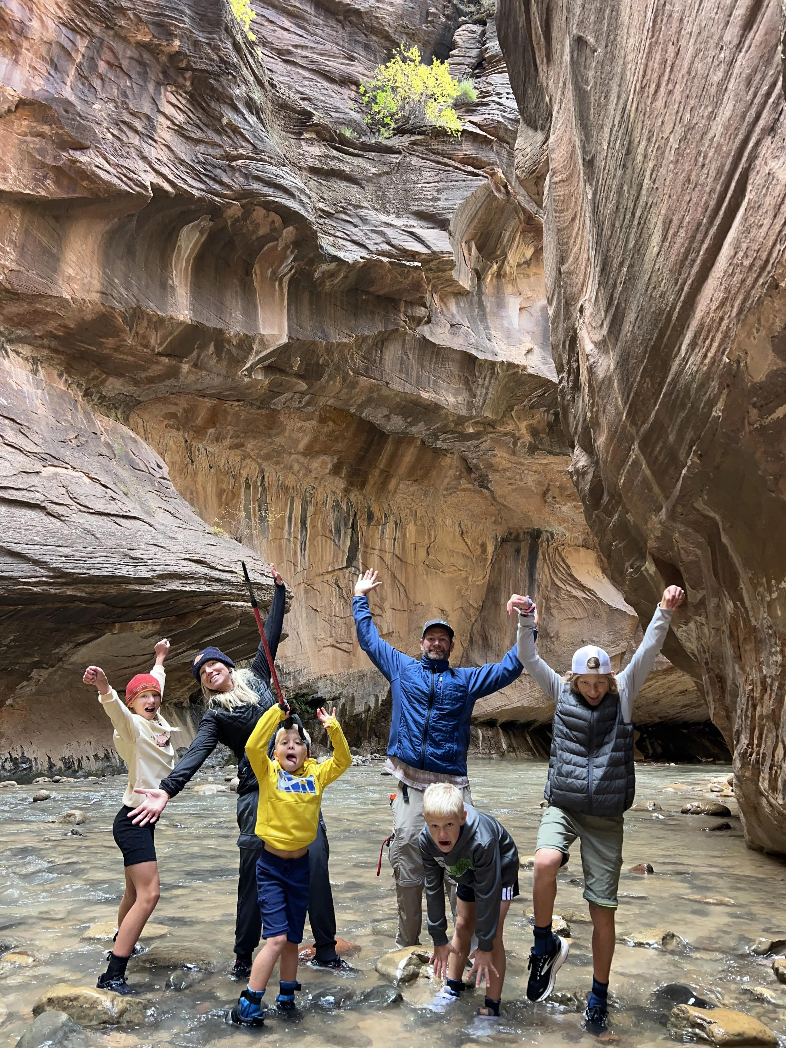 Group of seven people, including children and adults, standing and posing playfully in a shallow river inside a narrow canyon with steep, layered rock walls.