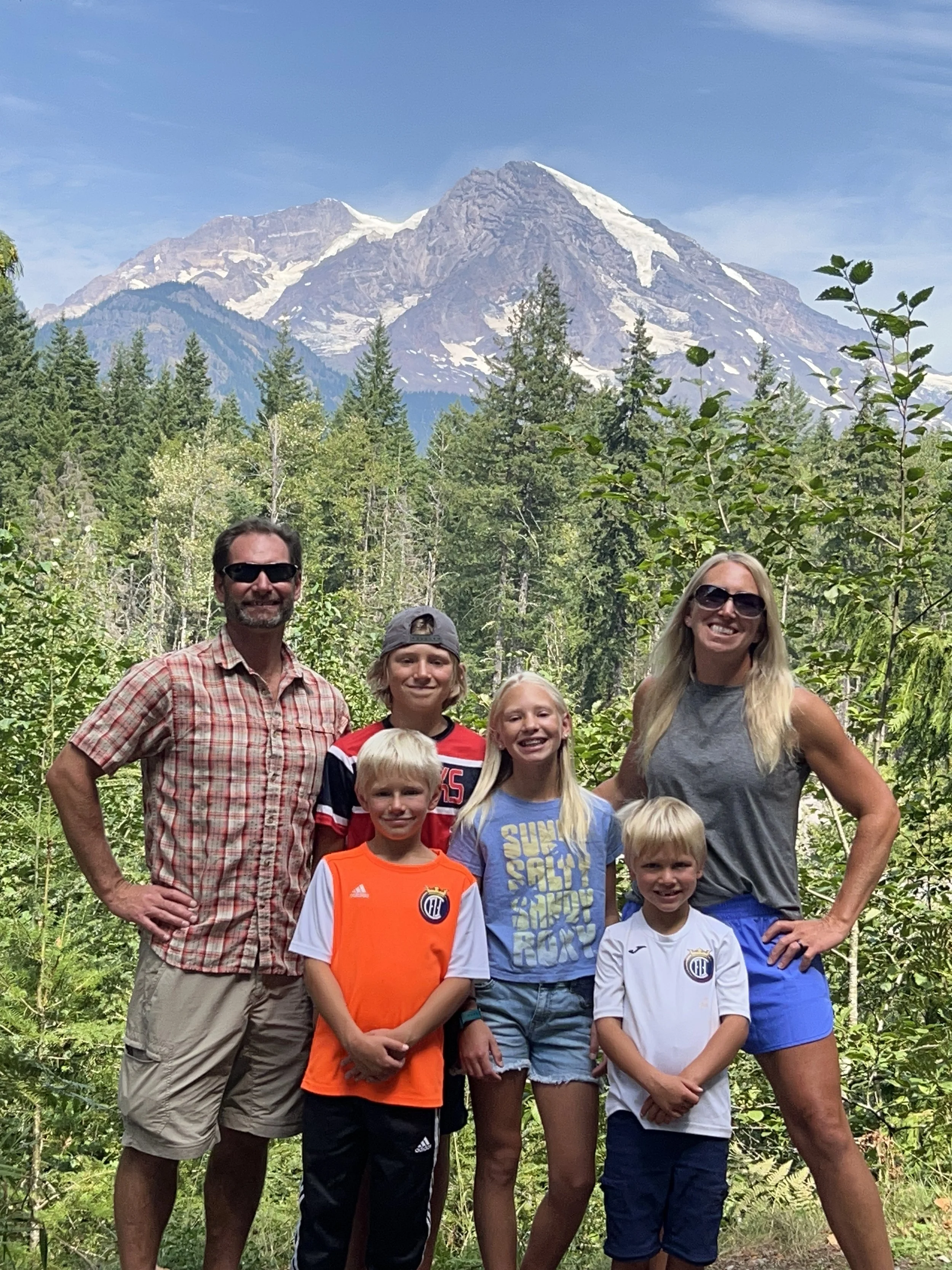 A family of six standing outdoors in a forest with Mt. Rainier in the background. They are smiling and wearing casual clothing, some with sunglasses.
