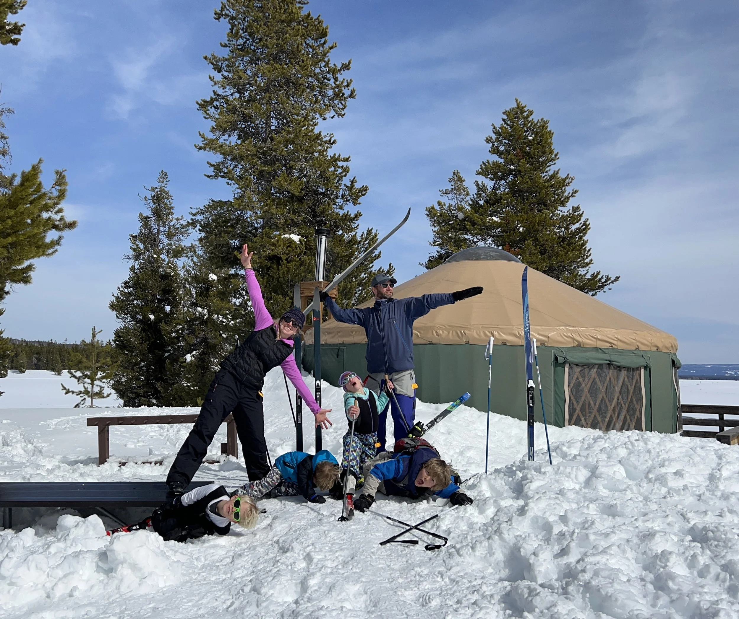 A group of six people, including children, are celebrating outside in the snow near a large tent and pine trees, with some lying on the snow and others standing and posing with ski equipment.