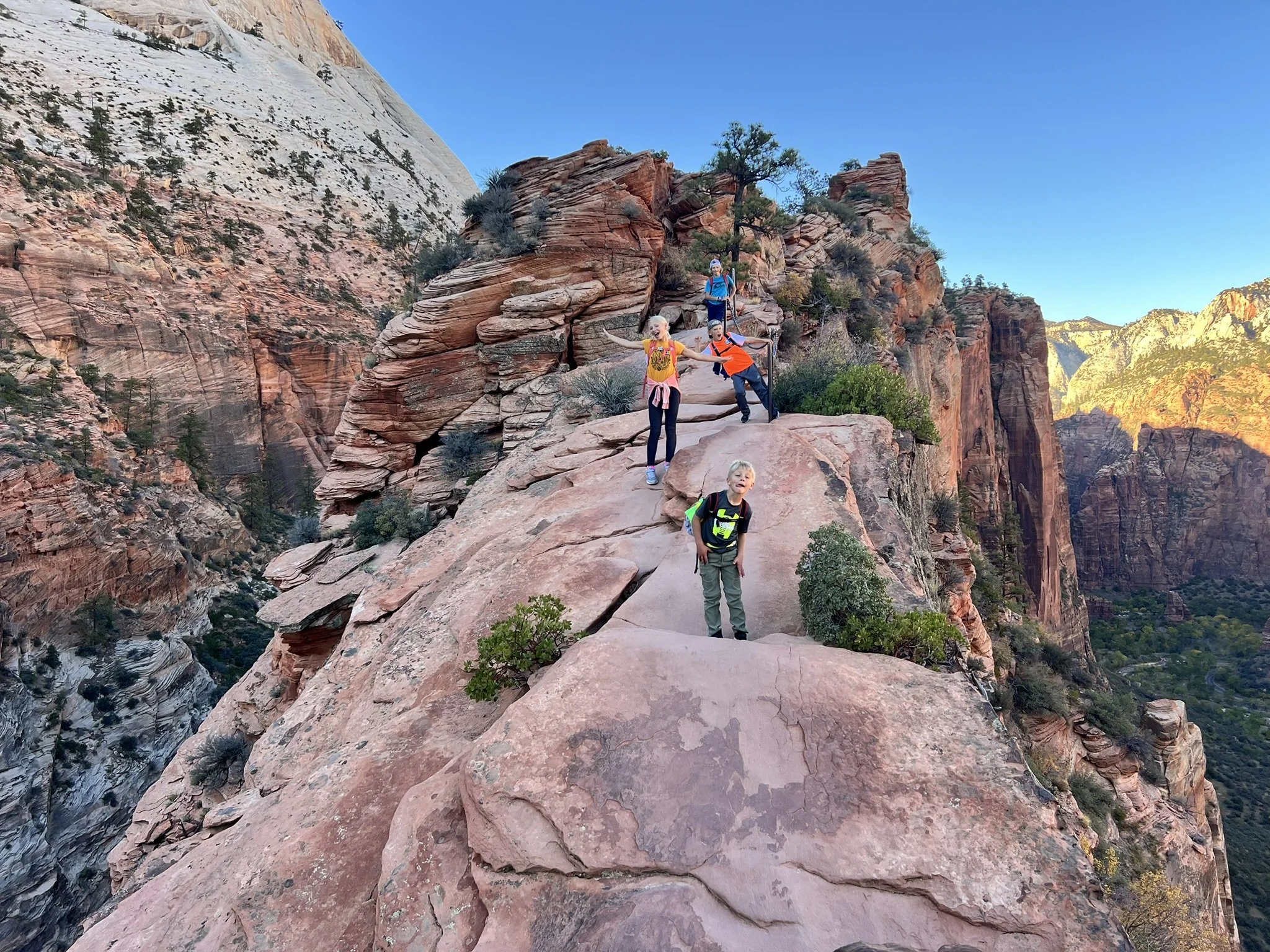 Group of children hiking on a rocky trail along a cliff in a desert canyon landscape during daytime.