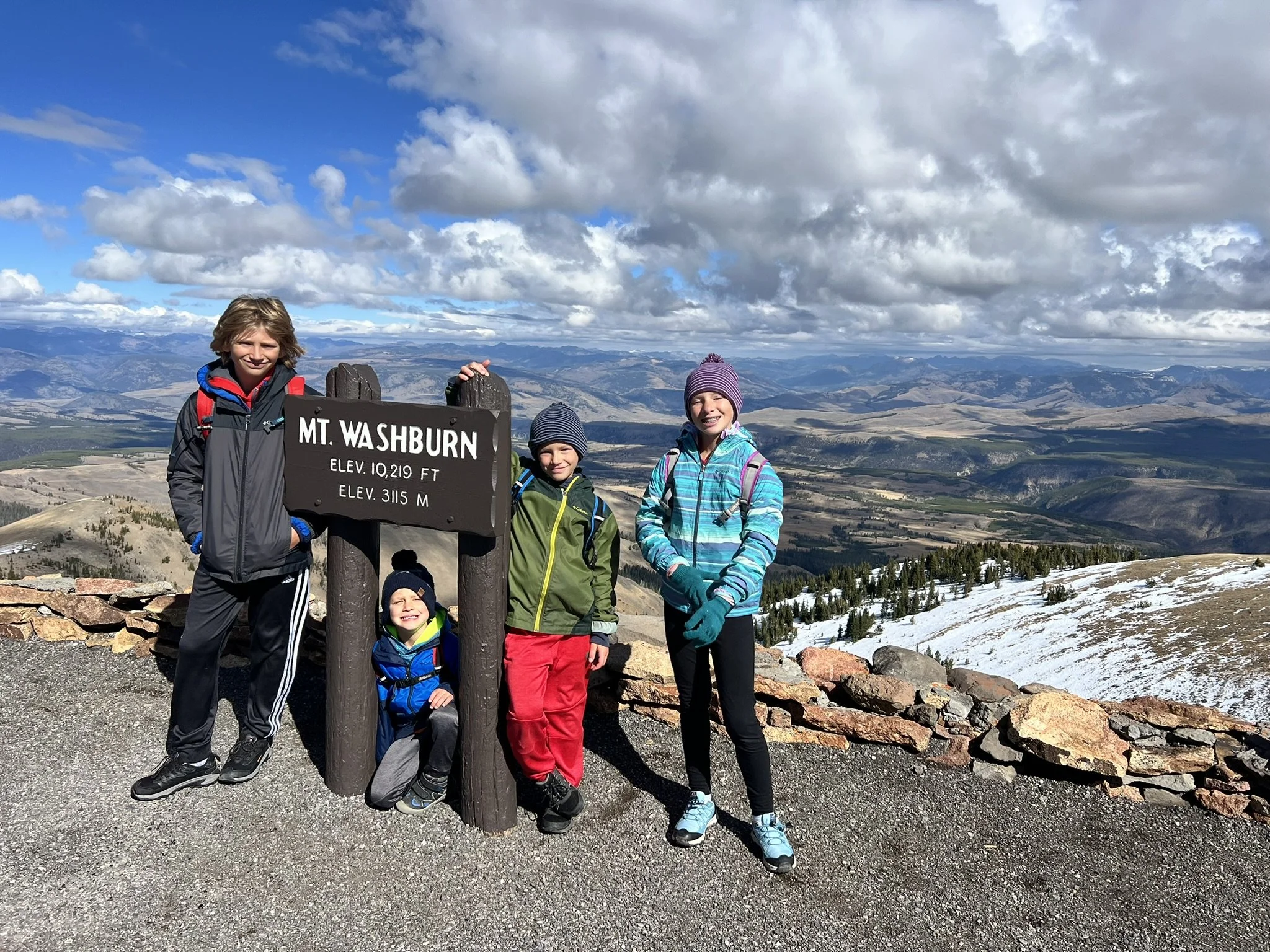 Four children standing next to a sign that reads 'Mt. Washburn, Elev. 10,219 ft, Elev. 3115 m,' on top of a mountain with a scenic landscape of valleys, mountains, and a partly cloudy sky in the background.