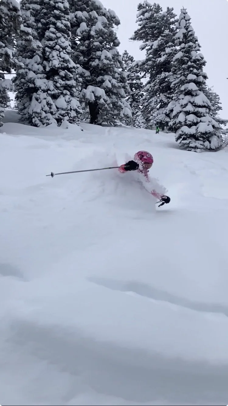A young girl in a pink helmet and pink jacket falling or sliding in deep snow with tall snow-covered pine trees in the background.