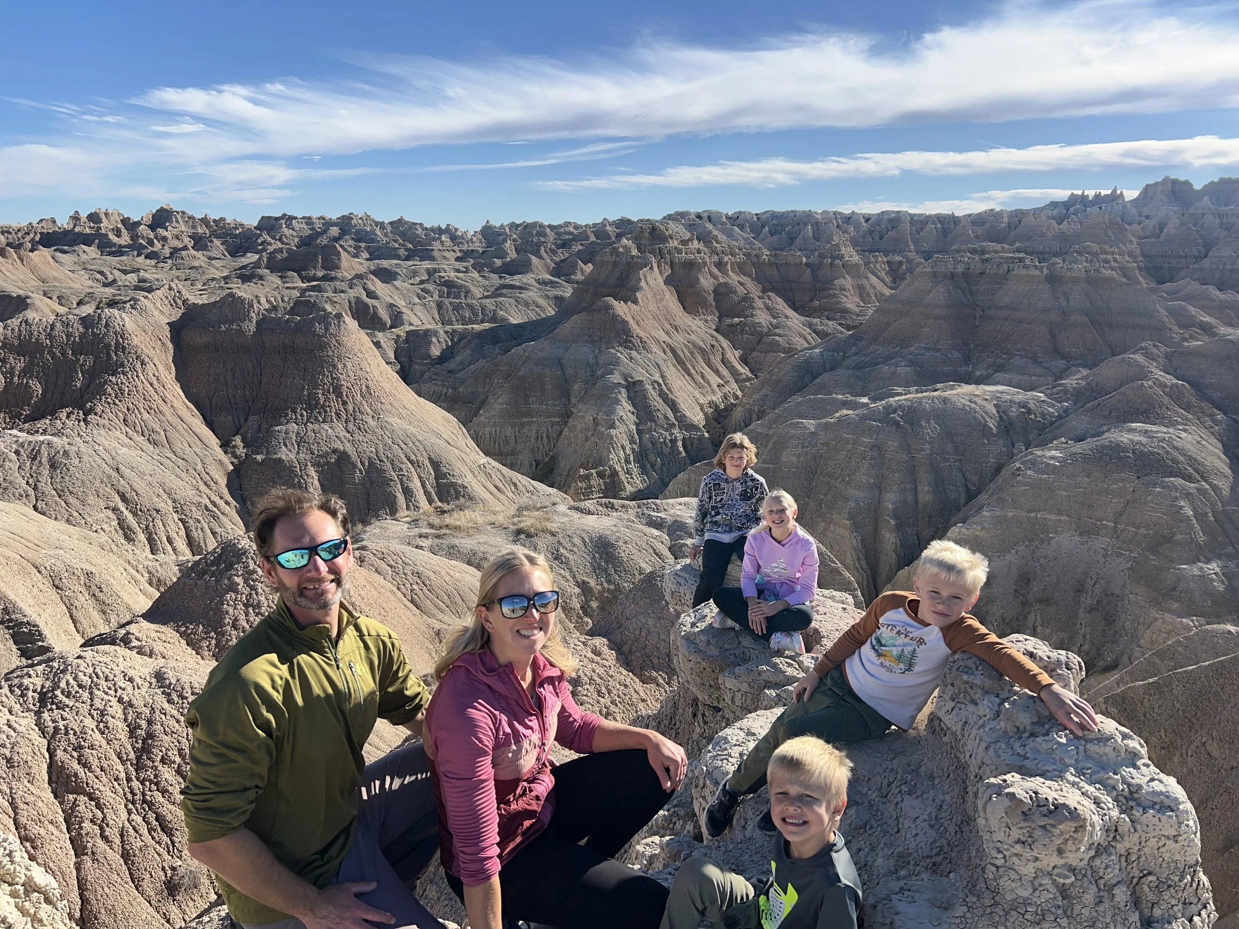A family of six smiling and posing on rocks with the Badlands in the background under a partly cloudy sky.