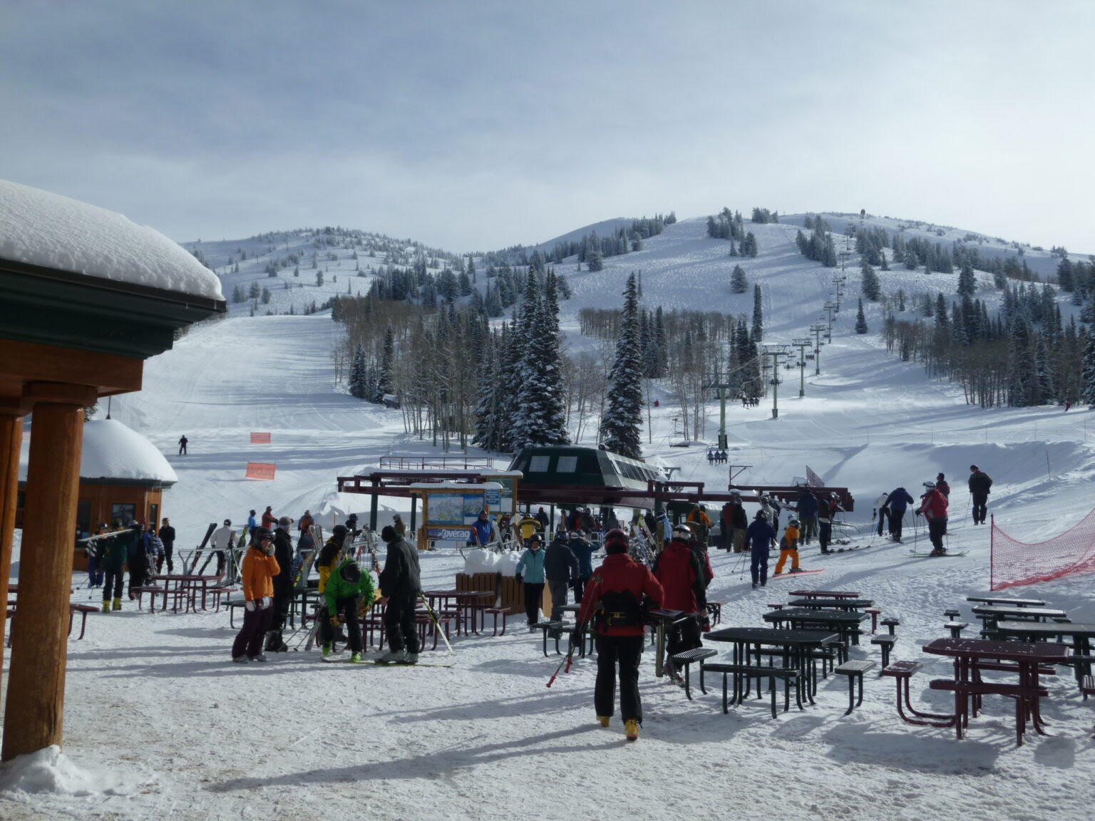 Ski resort with people in winter clothing, ski equipment, and tables outside, surrounded by snow-covered trees and mountains, ski lifts in the background.