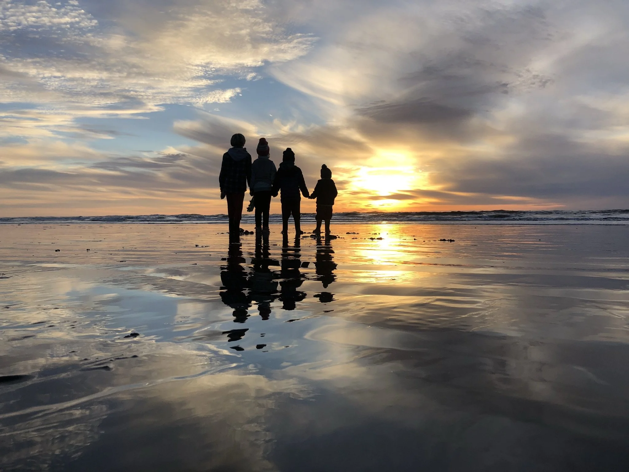 Four people holding hands walking on wet sand at the beach during sunset with clouds in the sky.