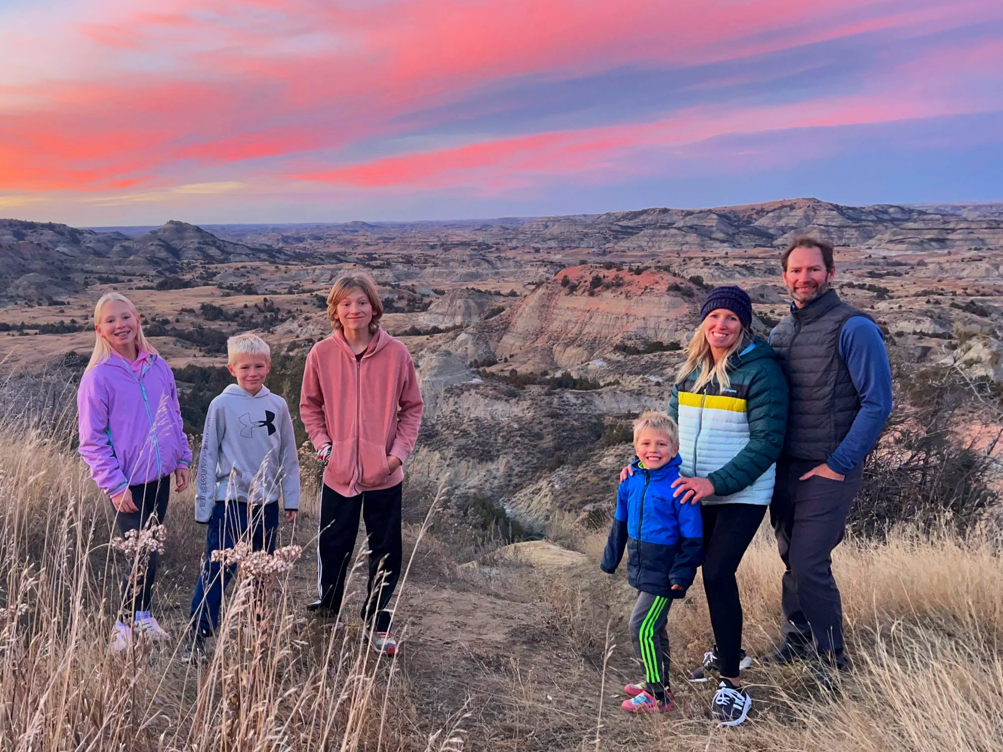 Family of six standing on a trail in a hilly desert landscape at sunset, smiling, dressed in colorful jackets and athletic clothing.
