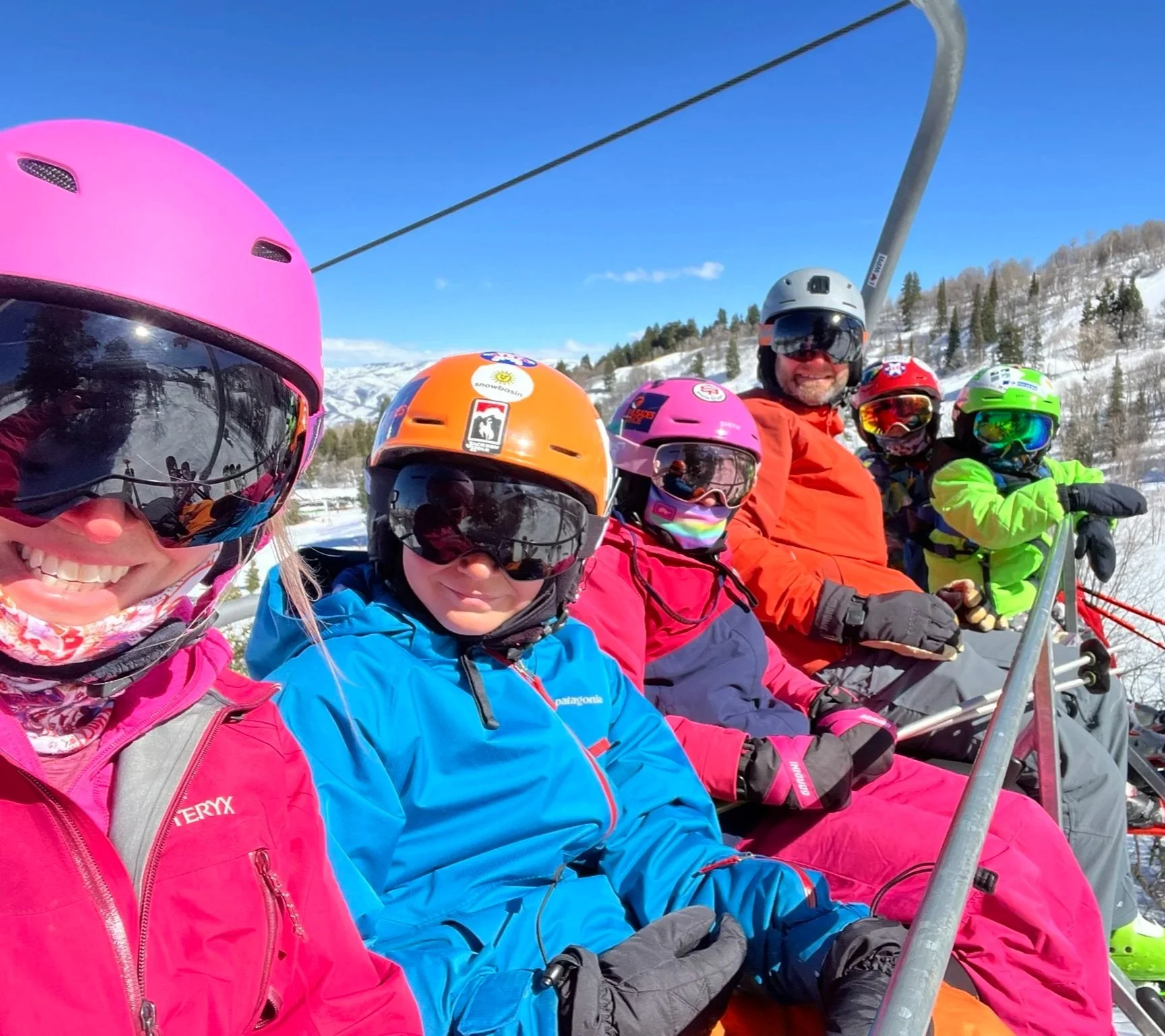 Group of skiers sitting on a ski lift, dressed in colorful ski gear with helmets and goggles, on a snowy mountain.