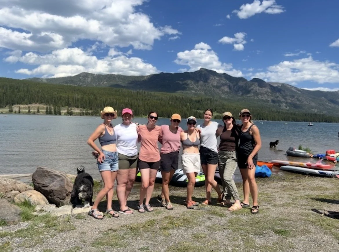 Group of eight women in summer clothing standing together near a lake with mountains in the background, some wearing sunglasses and hats, next to paddleboards and a dog.