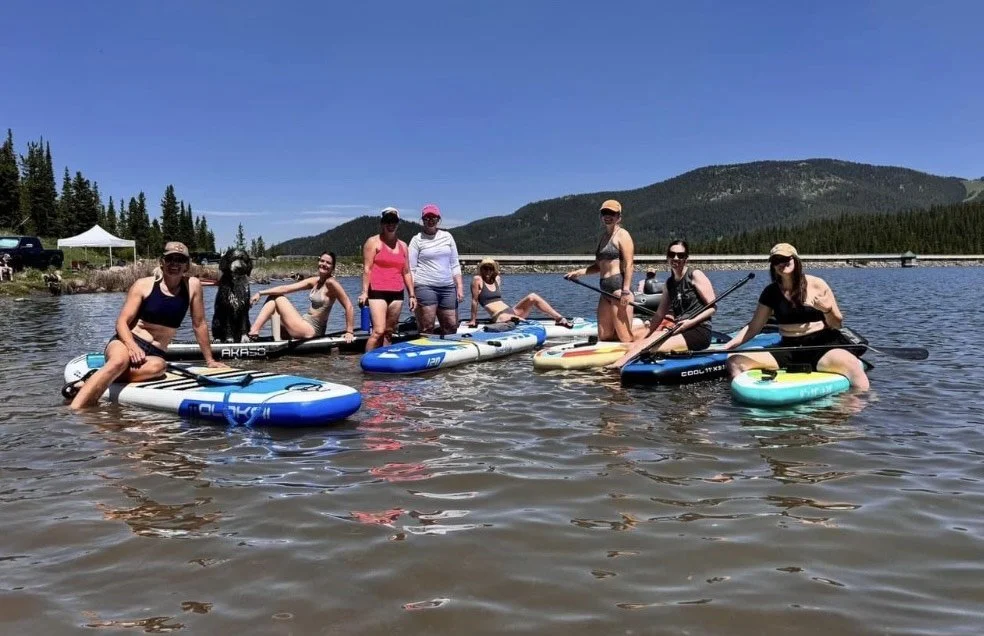 Group of people on paddleboards and sitting in shallow water at a lake, with mountains and trees in the background on a sunny day.