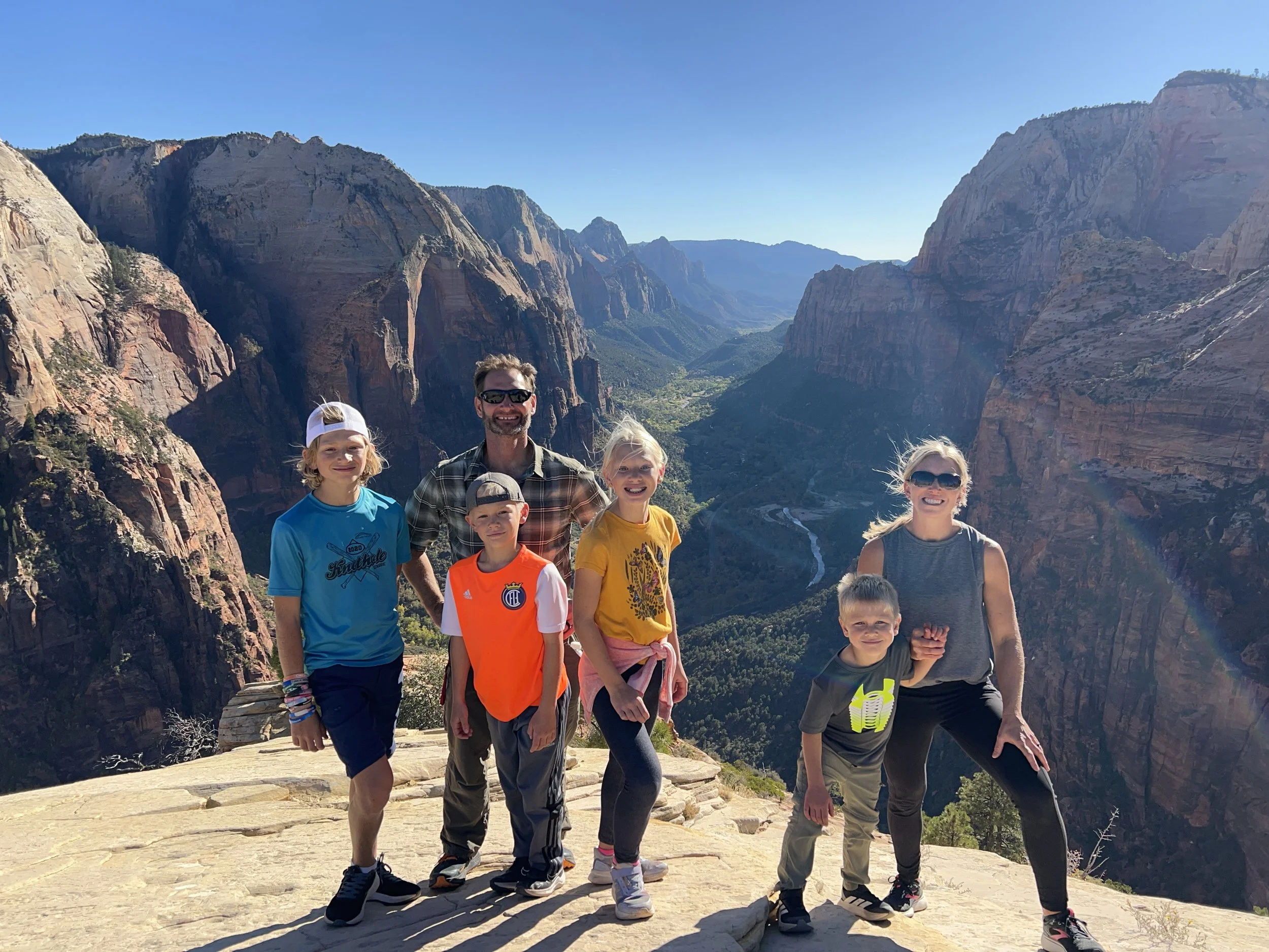 Family of seven standing on a rocky ledge overlooking a canyon with steep cliffs and a winding river in Zion National Park on a sunny day. They are smiling and dressed in casual outdoor clothing.