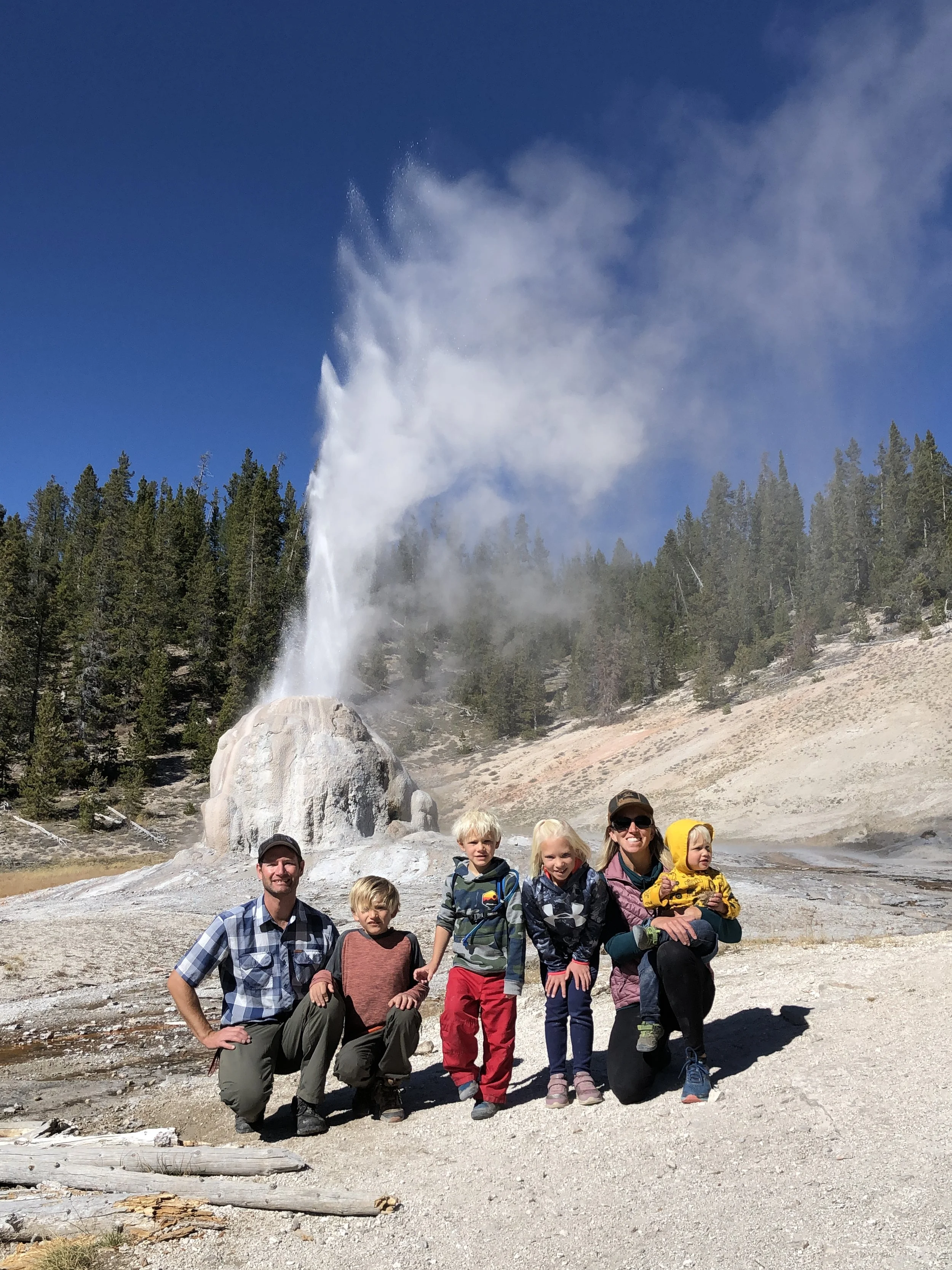A family of six posing in front of a geyser eruption at Yellowstone National Park on a sunny day.