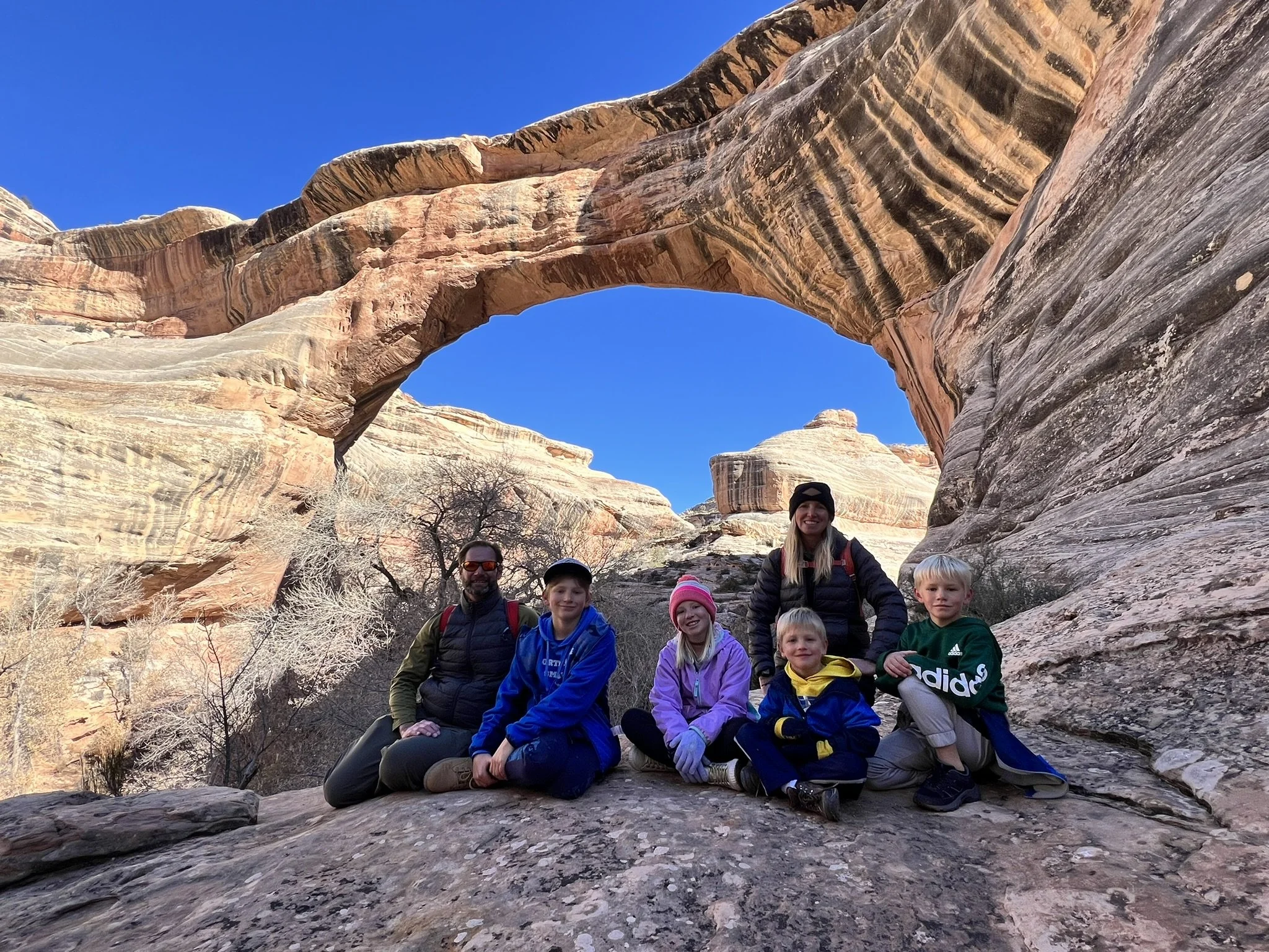 A group of six people, including two adults and four children, sitting and kneeling on a large rock in front of a natural rock arch formation in a desert landscape with clear blue sky and some trees in the background.