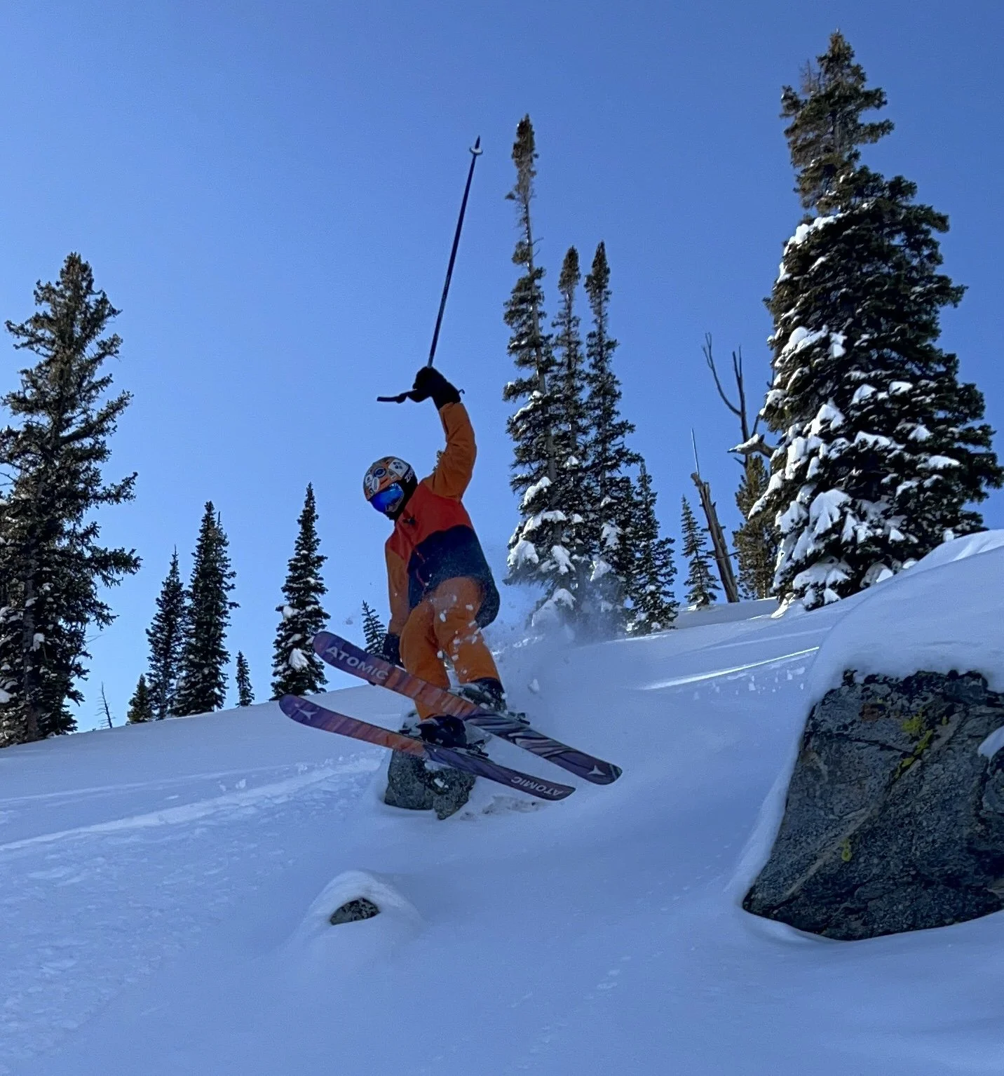 A skier in colorful gear jumping off a snow-covered rock in a forested mountainous area, with snow-laden pine trees and a clear blue sky in the background.