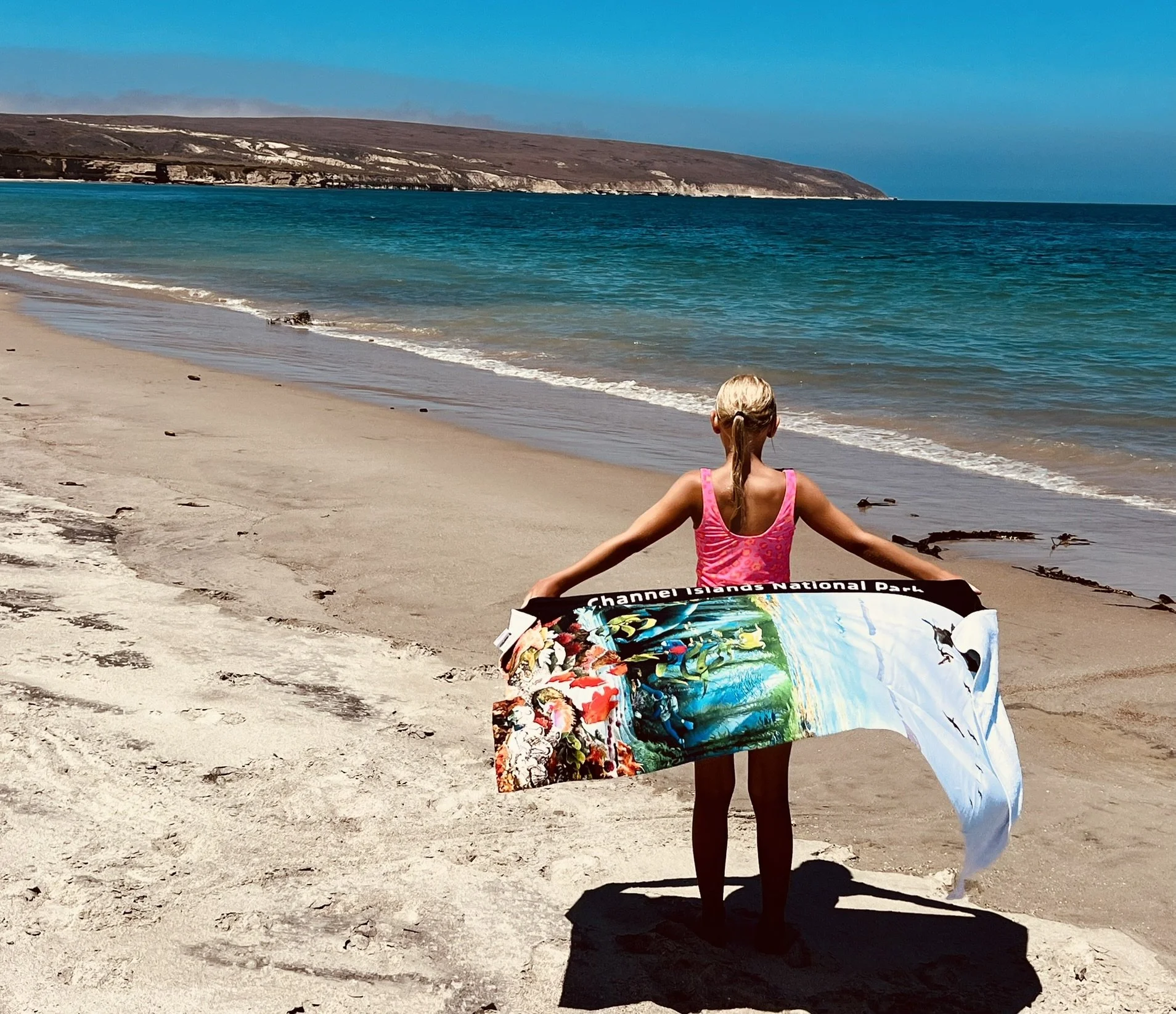 A young girl in a pink swimsuit holding a colorful towel, standing on a sandy beach facing the ocean, with cliffs in the distance and a clear blue sky.