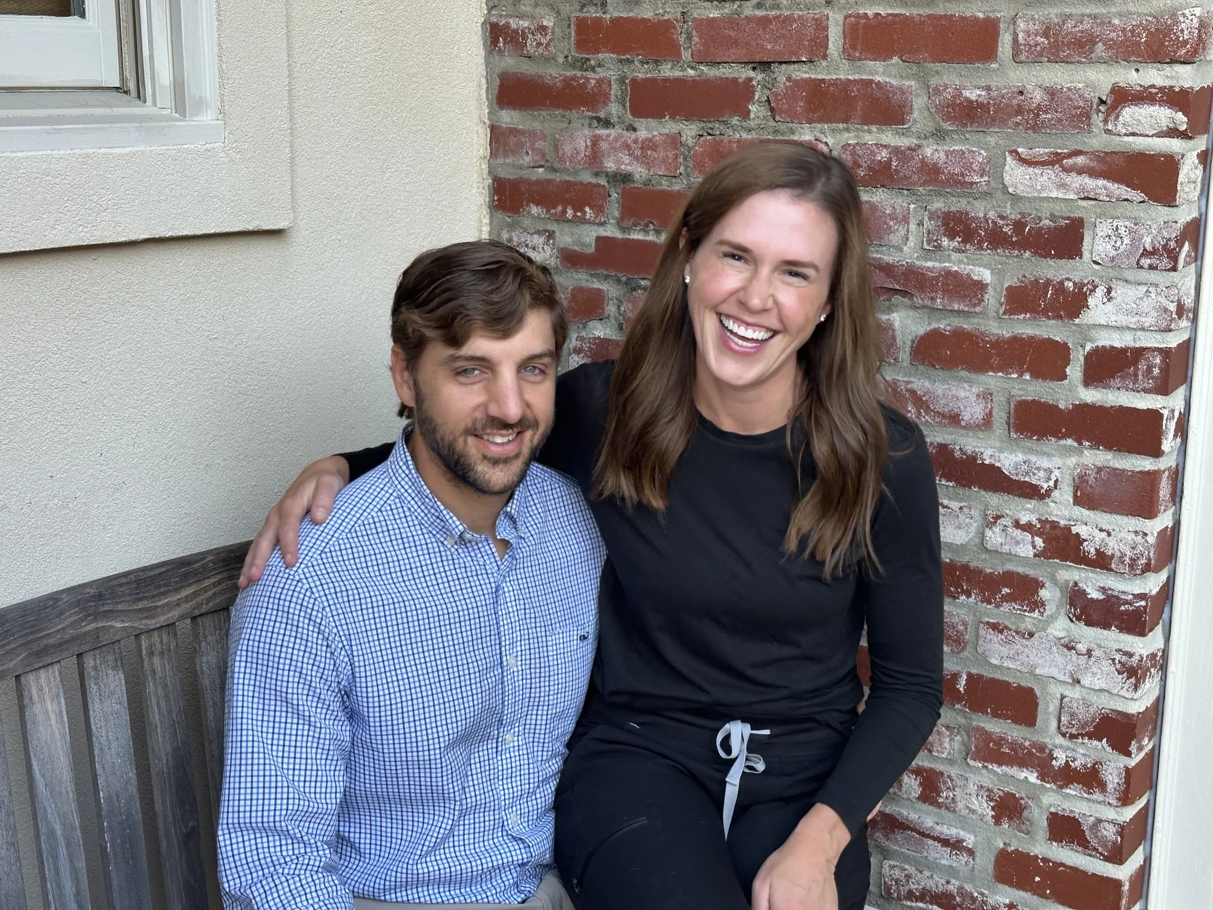 A man with brown hair and beard wearing a blue checkered shirt, and a woman with brown hair in a black long-sleeve shirt, sitting on a wooden bench with brick and beige wall backgrounds, smiling and posing for a photo.
