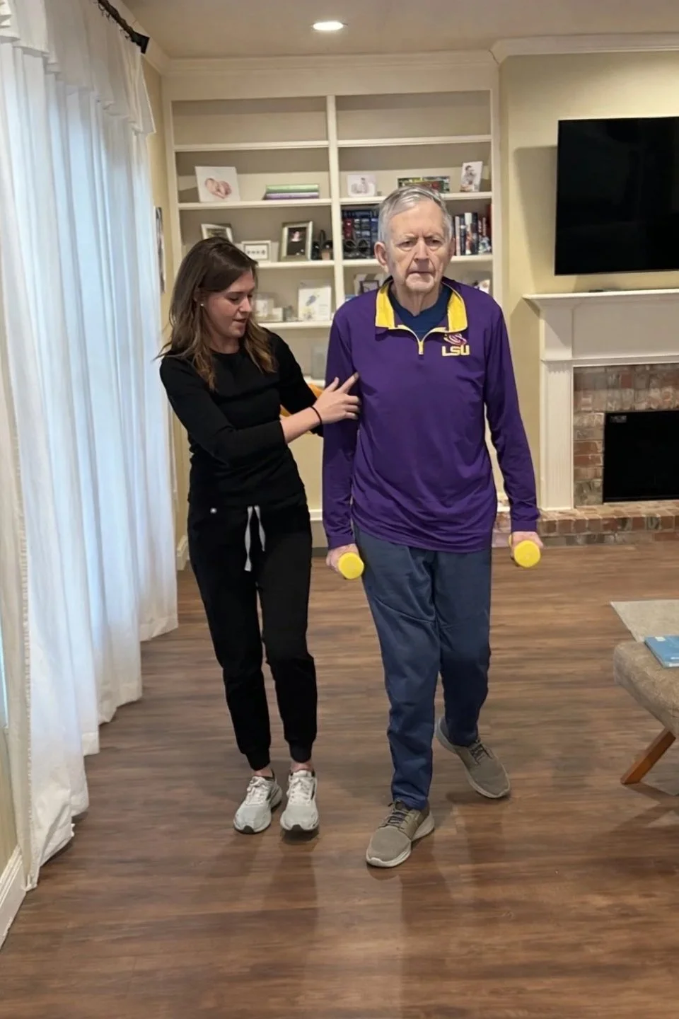 A young woman assists an elderly man with a mobility or physical therapy exercise indoors, holding yellow weights at his sides as he walks forward.