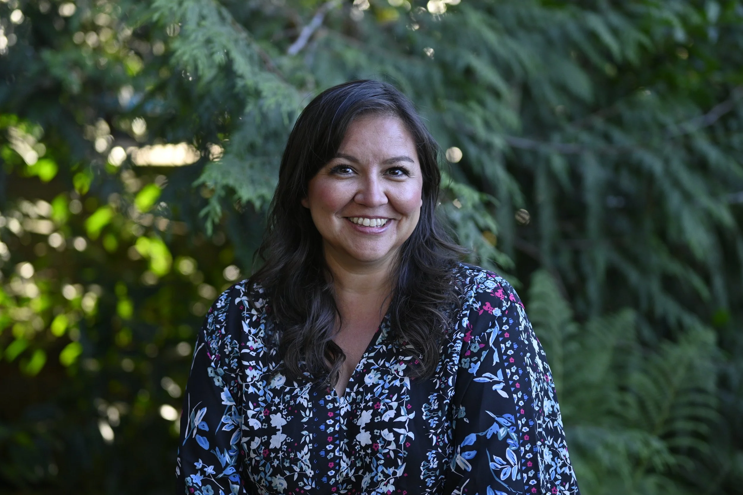 A woman with dark brown hair, smiling and wearing a black floral blouse, standing outdoors with green foliage in the background.