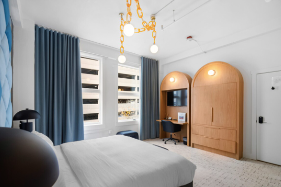 A modern bedroom featuring a bed with white linens, blue curtains, a wooden wardrobe, a small desk with a chair, a flat-screen TV, and a chandelier with yellow chains and white globes.