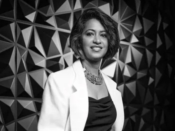 Black and white portrait of a woman with shoulder-length hair, wearing a white blazer over a black top, standing in front of a geometric patterned background.
