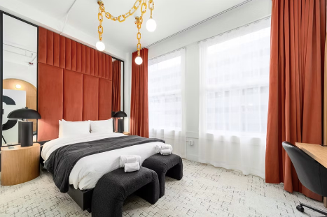 Modern hotel room with a bed, orange headboard, black bench at foot of bed, white bedding, black lamps, large windows with white curtains, orange drapes, and a desk with a black chair.