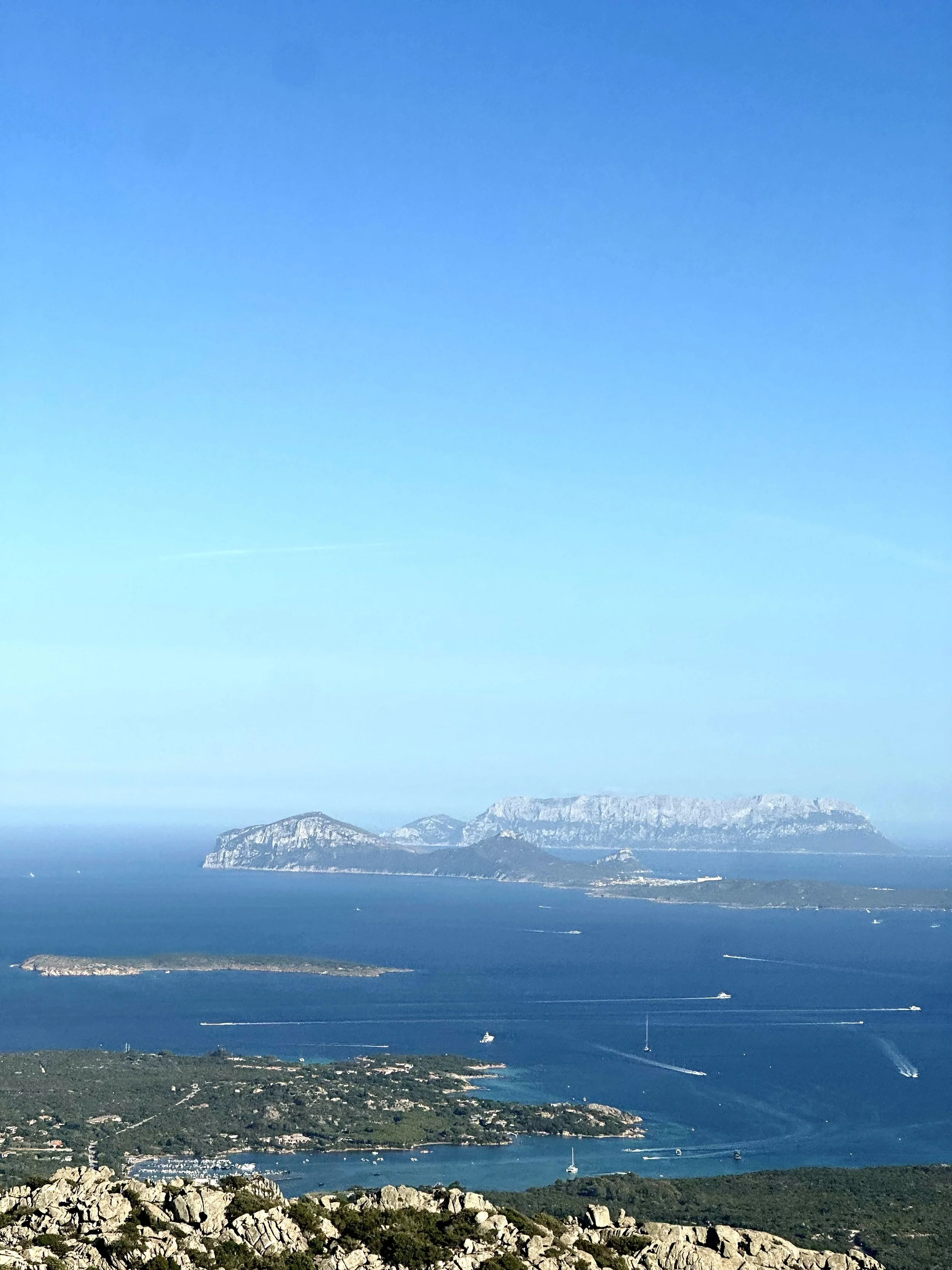 A panoramic view of a coastal landscape featuring a large island with mountainous terrain, surrounding smaller islands, a deep blue sea with numerous boats, and a rocky foreground under a clear blue sky.