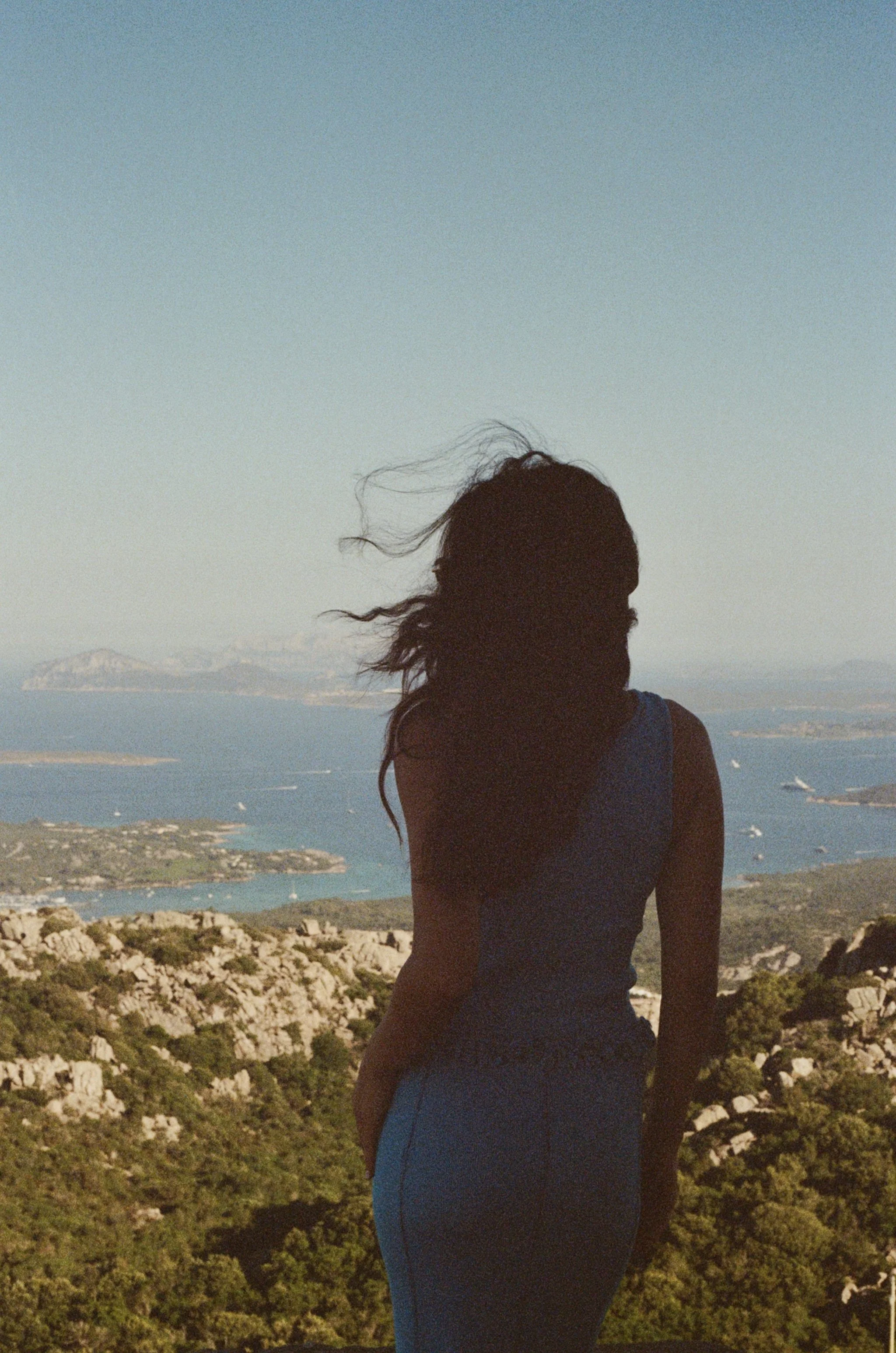 A person with long hair stands outdoors on a hill, overlooking a landscape with water and distant islands under a clear sky.