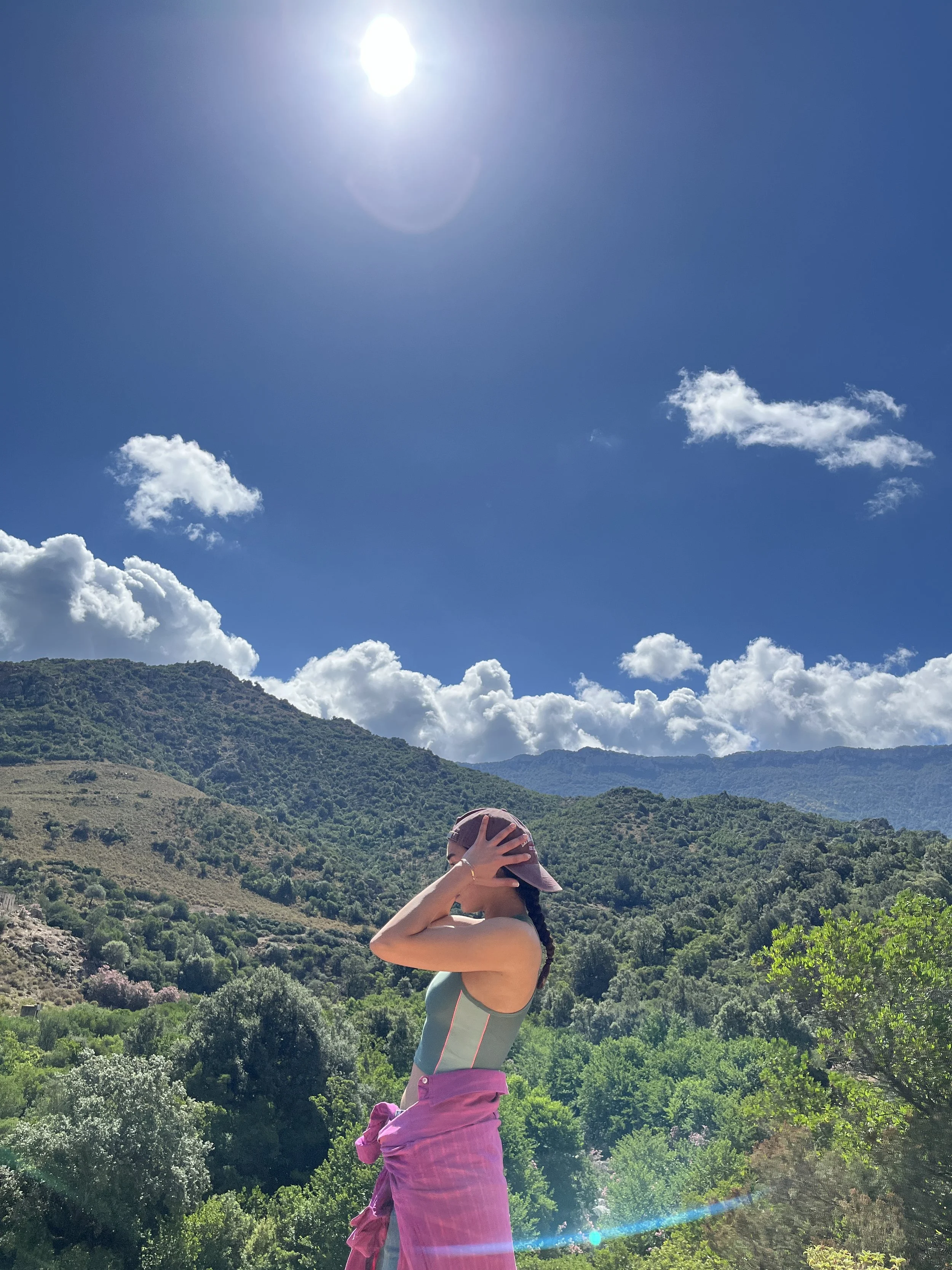 A woman standing in a lush green mountain landscape under a bright sunny sky with scattered clouds, wearing a pink hat and a pink jacket tied around her waist.