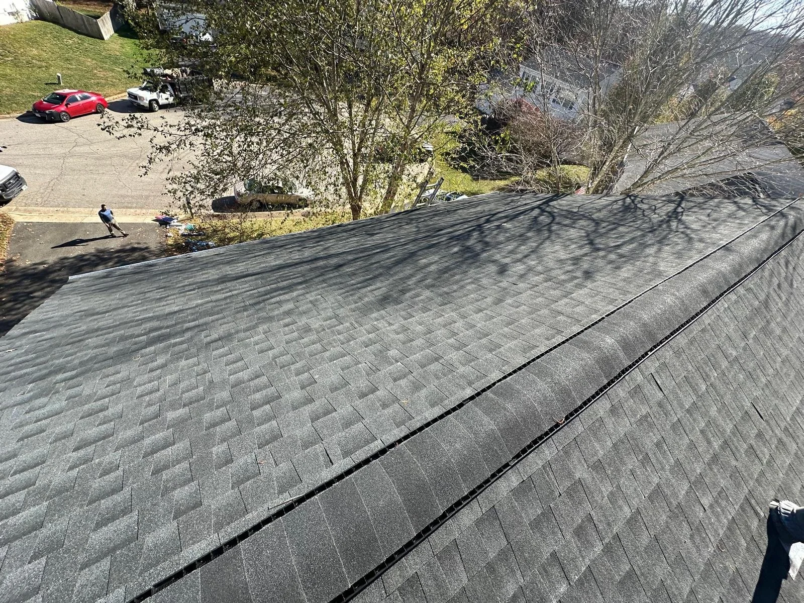 View of a residential roof with asphalt shingles, tree shadows, and neighboring cars and yard visible in the background.