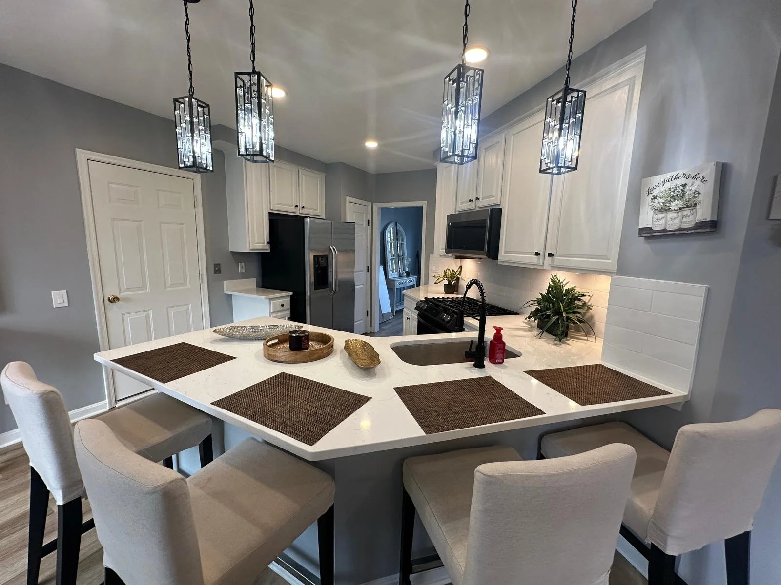 Modern kitchen with white cabinets, stainless steel refrigerator and oven, four pendant lights, and a breakfast bar with beige chairs.