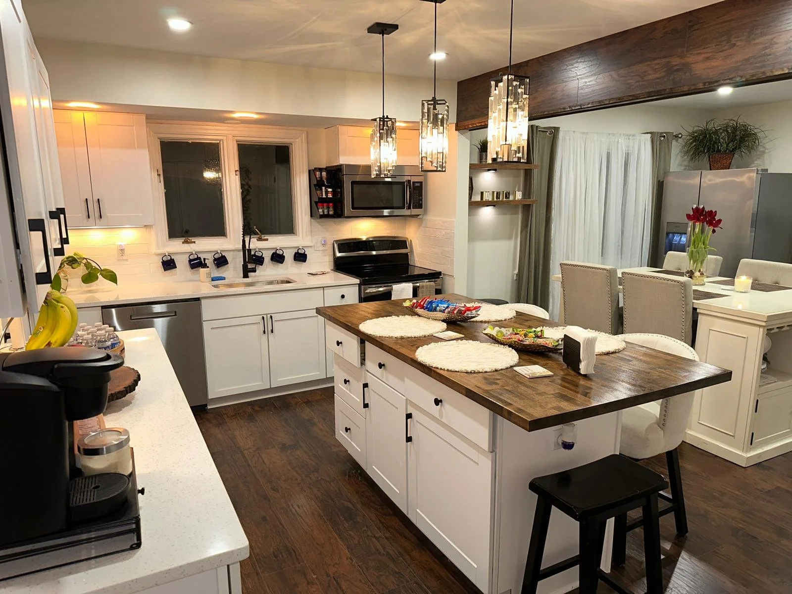 Modern kitchen with white cabinets, wooden island, stainless steel appliances, and a cozy dining area with beige chairs and a white table, decorated with a vase of red flowers and candles.