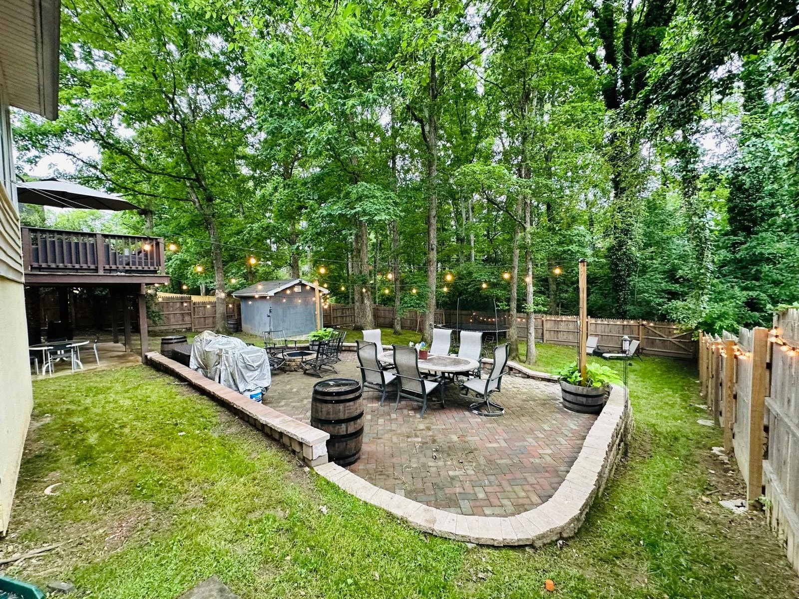 A backyard patio with outdoor furniture, string lights, and a small shed surrounded by trees and a wooden fence.