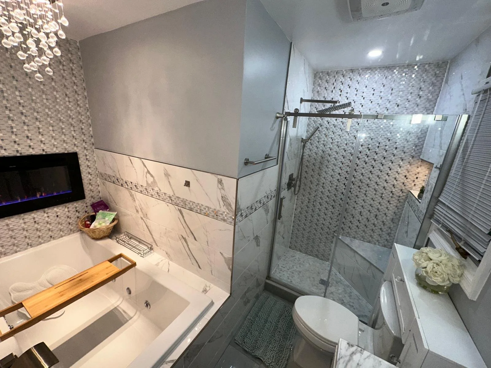 A modern bathroom featuring a walk-in shower with a glass enclosure, a wall-mounted fireplace, a bathtub with a wooden bath tray, a basket of toiletries, and a window with blinds. The decor includes marble and mosaic tiles.