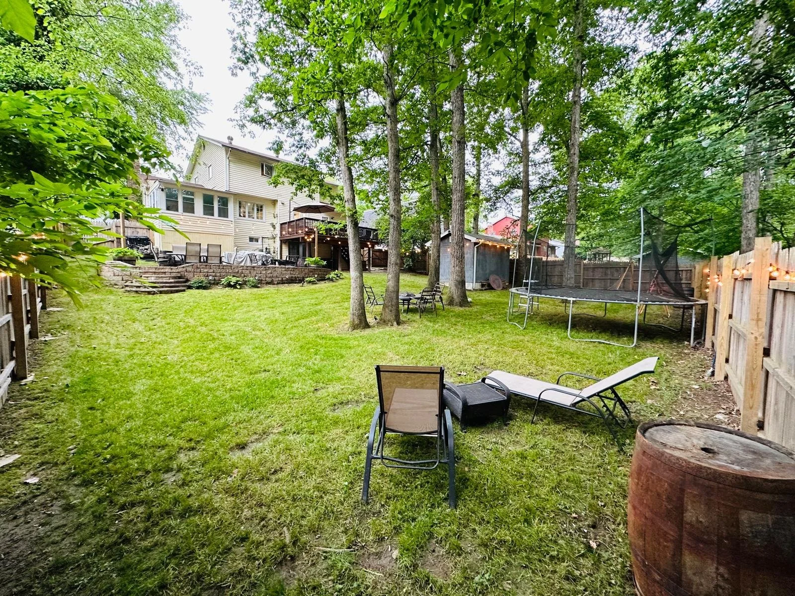 A backyard with green grass, several patio chairs, a trampoline, trees, a wooden fence, and a house with multiple decks and outdoor furniture.