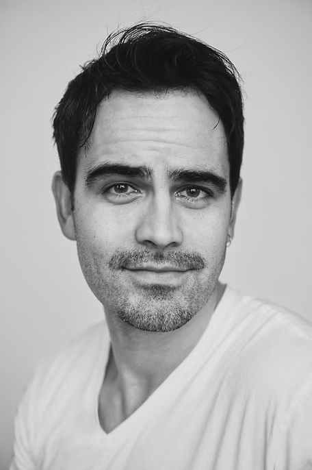 Black and white portrait of a man with short dark hair, slight stubble, wearing a white shirt, looking at the camera with a neutral expression.