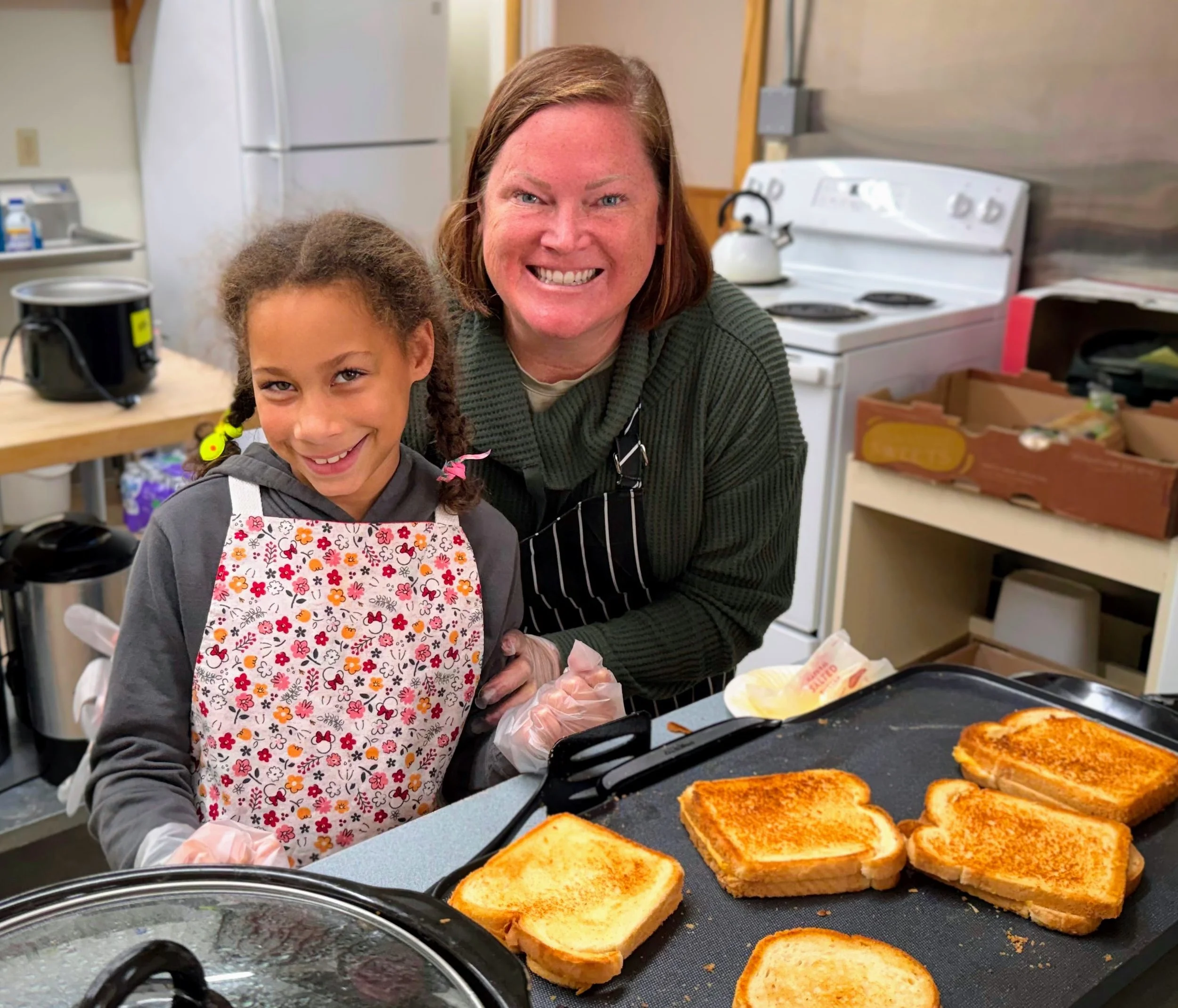 A woman and a girl preparing grilled cheese sandwiches in a kitchen, smiling at the camera. The girl is wearing a floral apron and the woman is wearing a striped apron, both standing behind a counter with toasted sandwiches on a grilling tray.