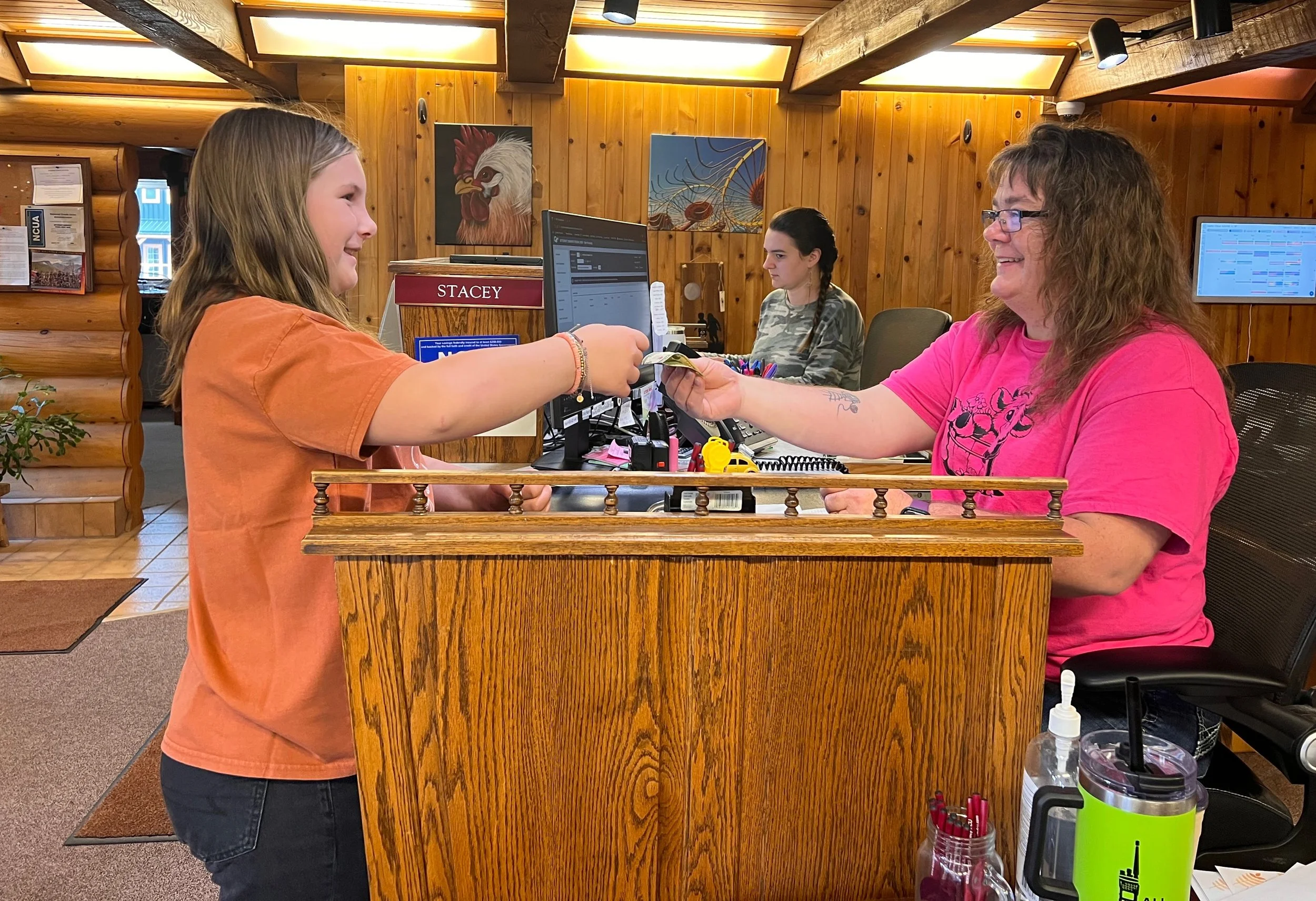 A young girl is at a reception desk handing money to a woman in a pink shirt, who is smiling. The woman is sitting at the desk in an indoor setting with wooden walls and artwork, with another woman working at a computer in the background.