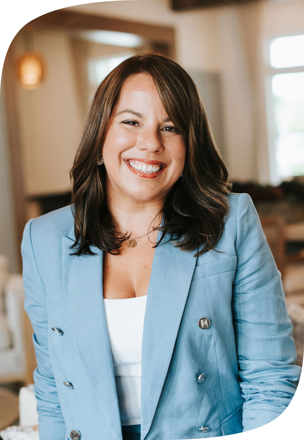 A woman with shoulder-length dark brown hair smiling, wearing a light blue blazer over a white top, with a blurred indoor background.