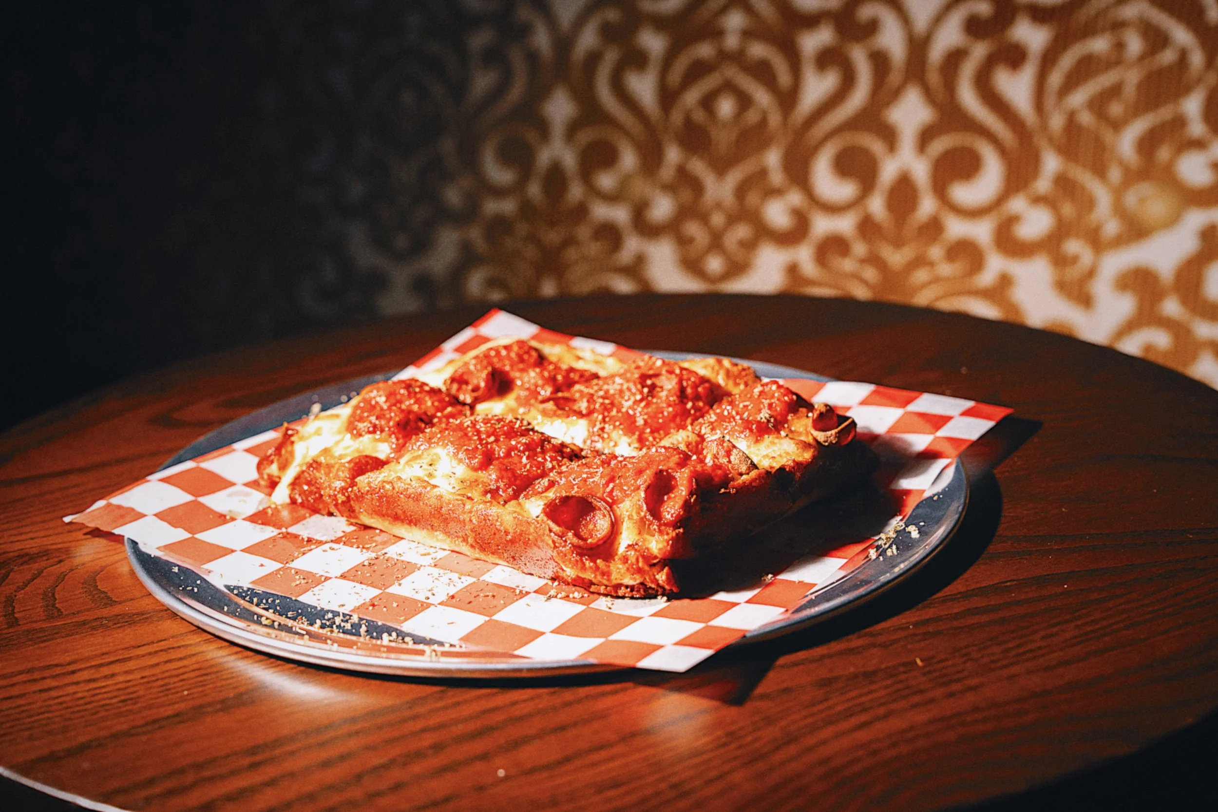 A slice of pepperoni pizza on checkered paper on a plate on a wooden table with a dark patterned wall background.