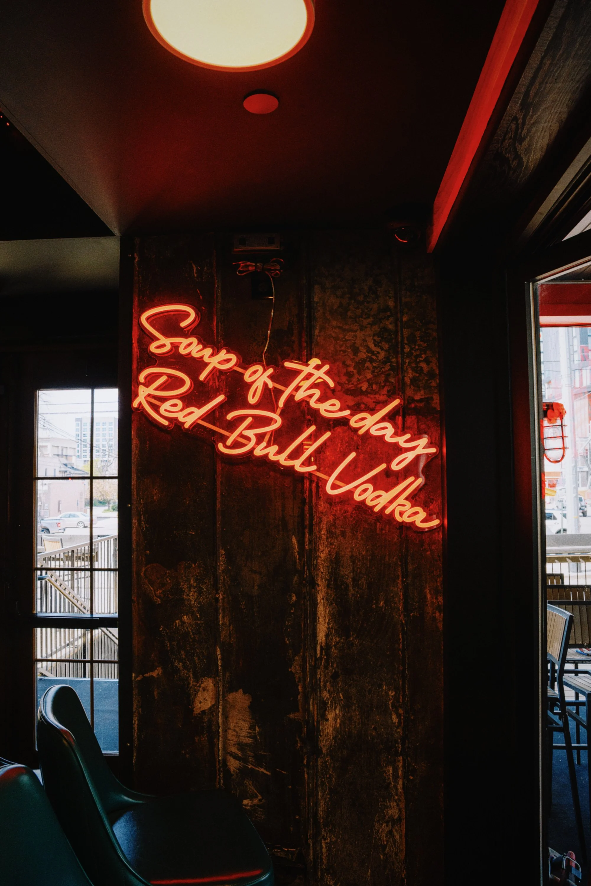 Neon sign in a restaurant reads 'Soup of the day Red Bull Vodka' against a wooden wall, with a window and outdoor seating visible.