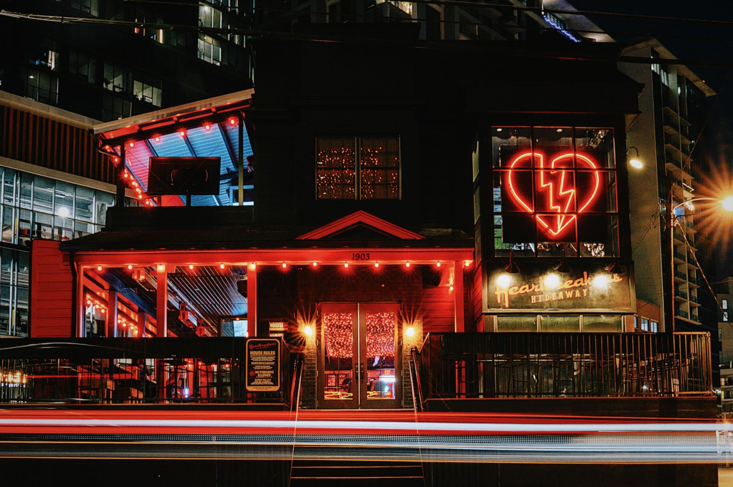 Nighttime view of a modern building with red neon signs, including a broken heart with a lightning bolt, and a lit-up entrance with cars passing by.
