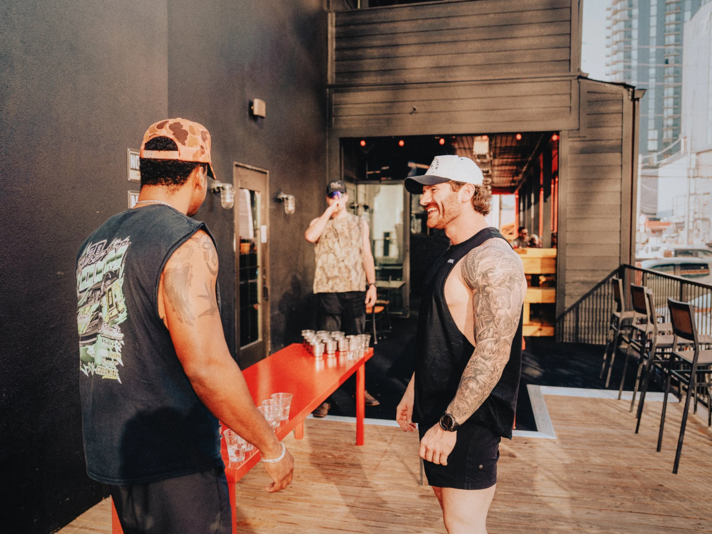 Two men with tattoos wearing sleeveless shirts and caps laughing and talking outside a building, with another man in the background drinking from a glass, blue sky, and city buildings in the distance.