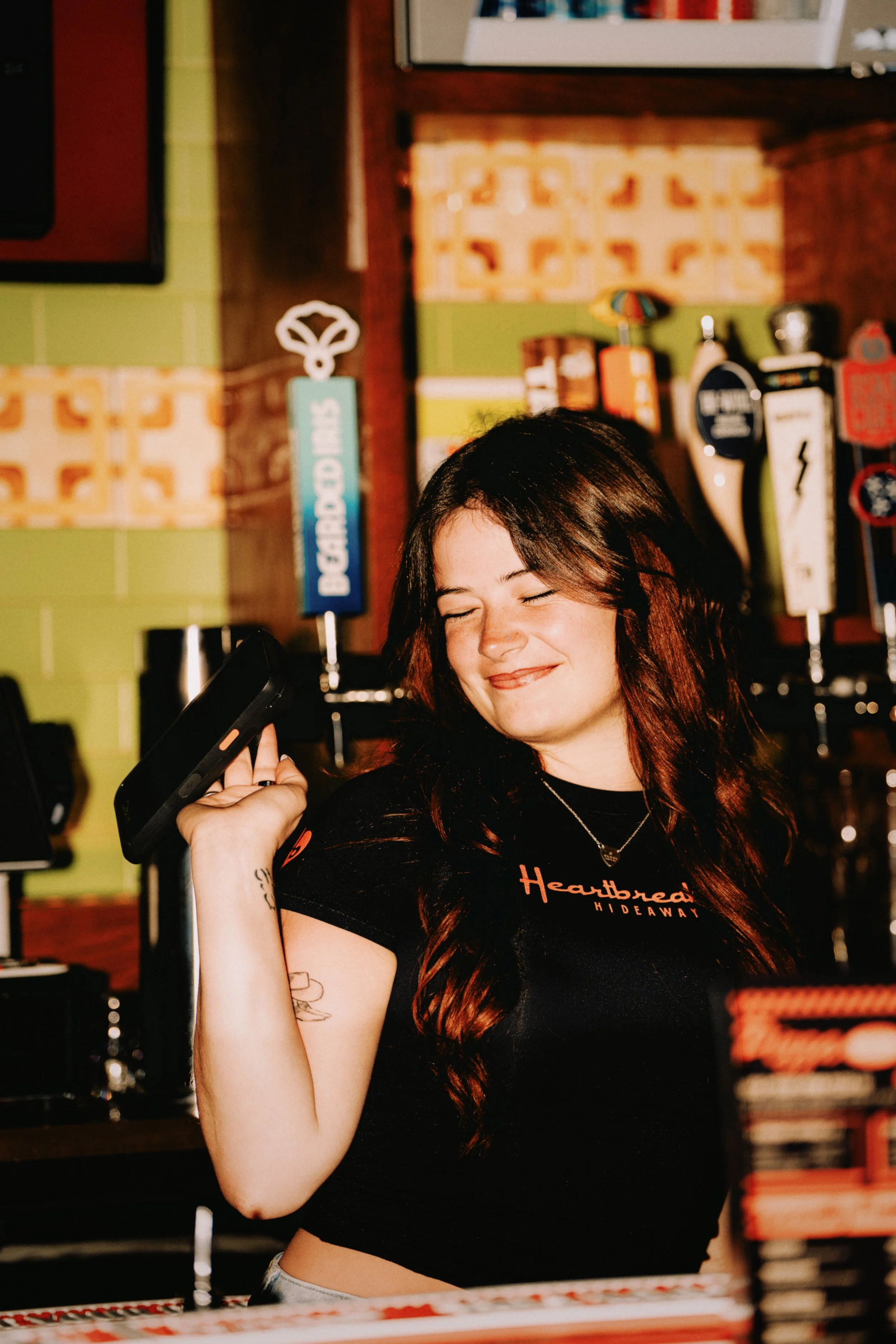 Young woman with long reddish-brown hair smiling with eyes closed, holding a phone on her shoulder in a bar or pub with colorful signs and beer taps in the background.