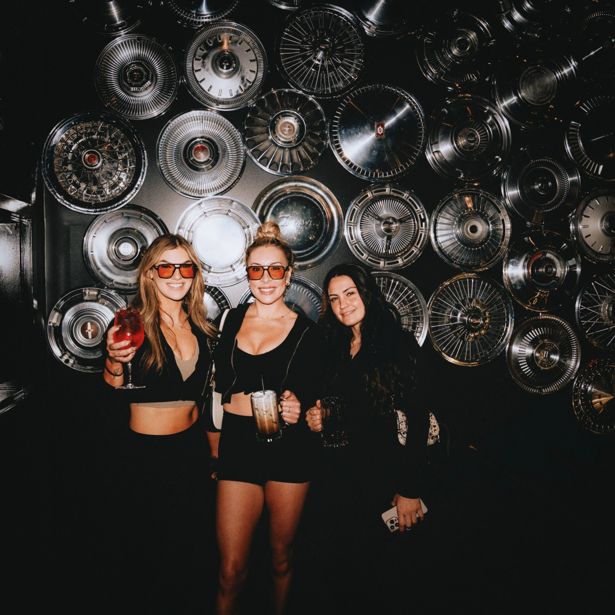 Three women smiling and holding drinks in front of a wall with vintage car hubcaps.