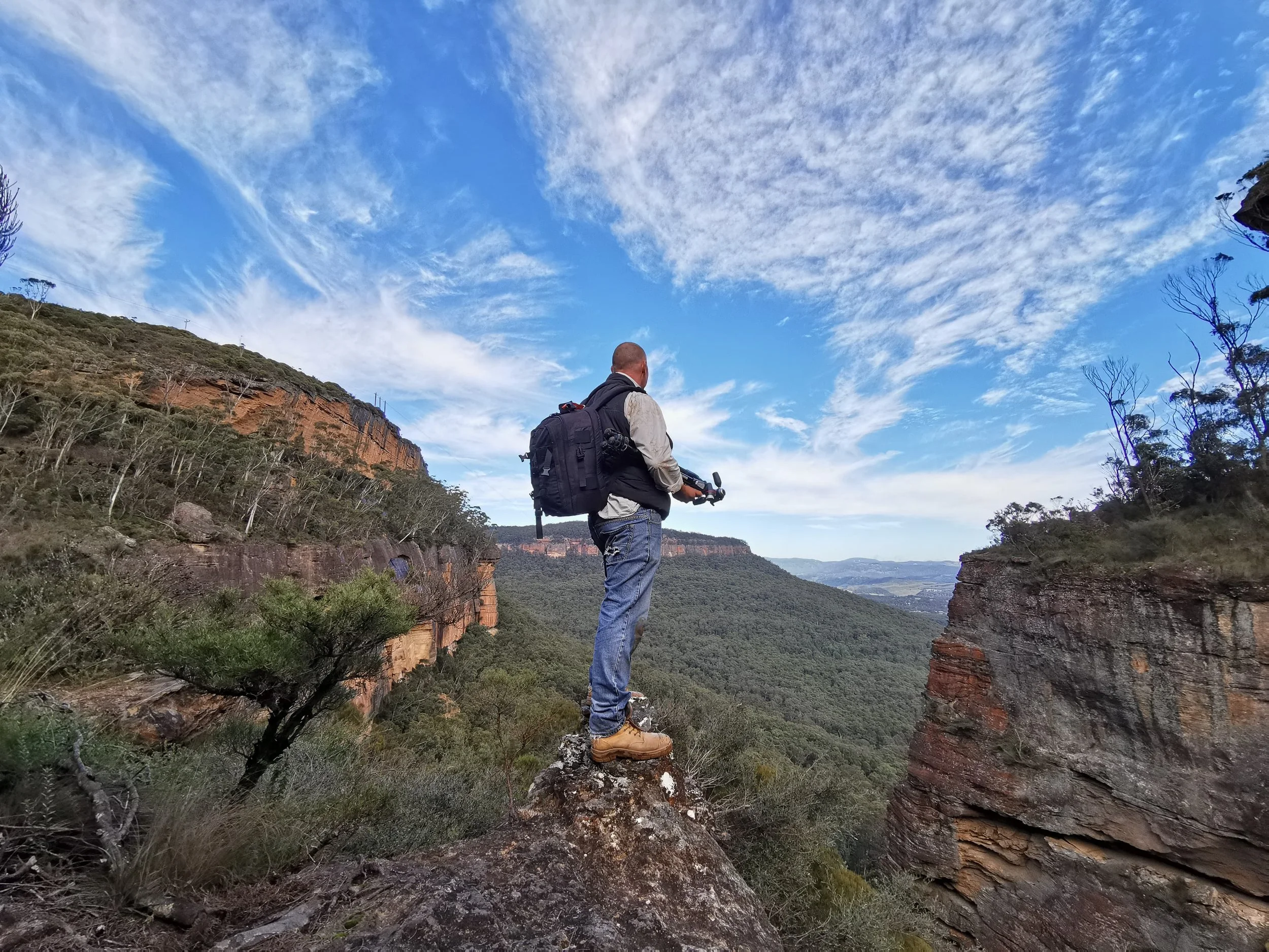 Jeff Walsh stands on a rock ledge overlooking a canyon with dense forest, holding a drone controller, under a mostly cloudy blue sky.