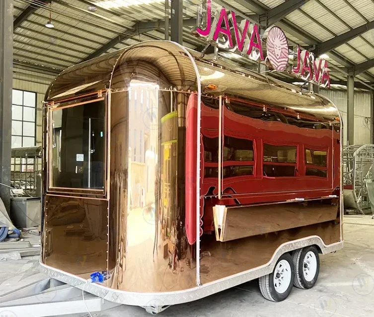 A vintage-style food cart with a shiny, reflective copper exterior, featuring a red windowed section and a sign on top that reads "JAVA JAVA" with a coffee bean graphic, inside a warehouse.