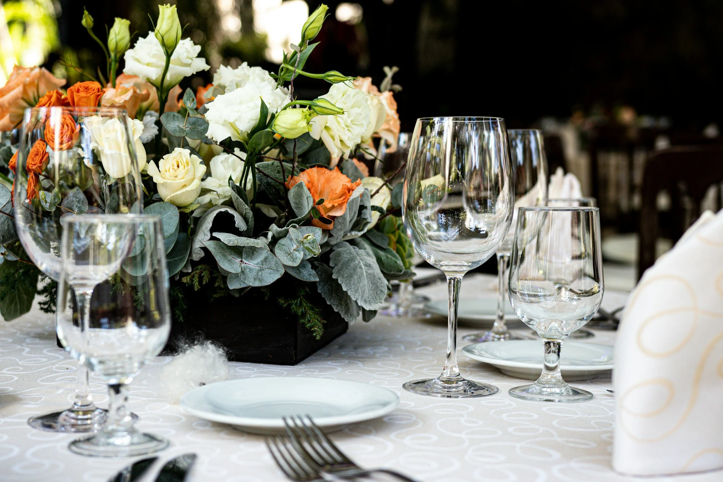 Set table with glassware, plates, silverware, and a floral centerpiece with white, cream, and orange flowers on a white tablecloth.