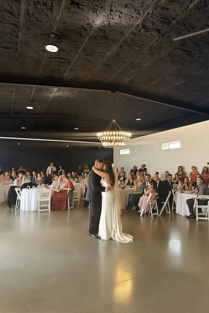 A bride and groom sharing a first dance at their wedding reception in a large, modern reception hall with black and white walls, a chandelier, and seated guests watching the couple.