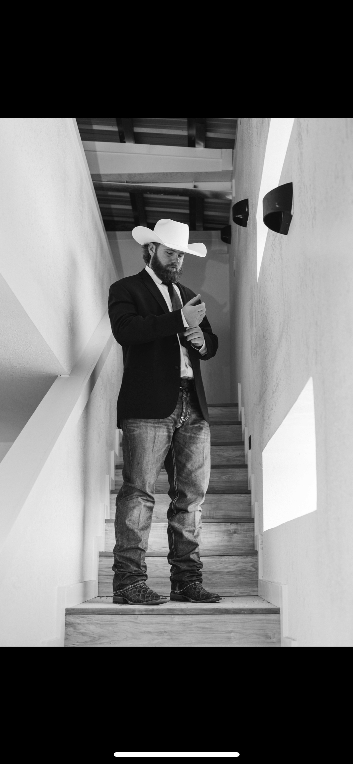 A man dressed in a blazer, jeans, cowboy boots, and a cowboy hat is standing on a staircase, looking at his phone in a modern interior with black speakers on the wall.