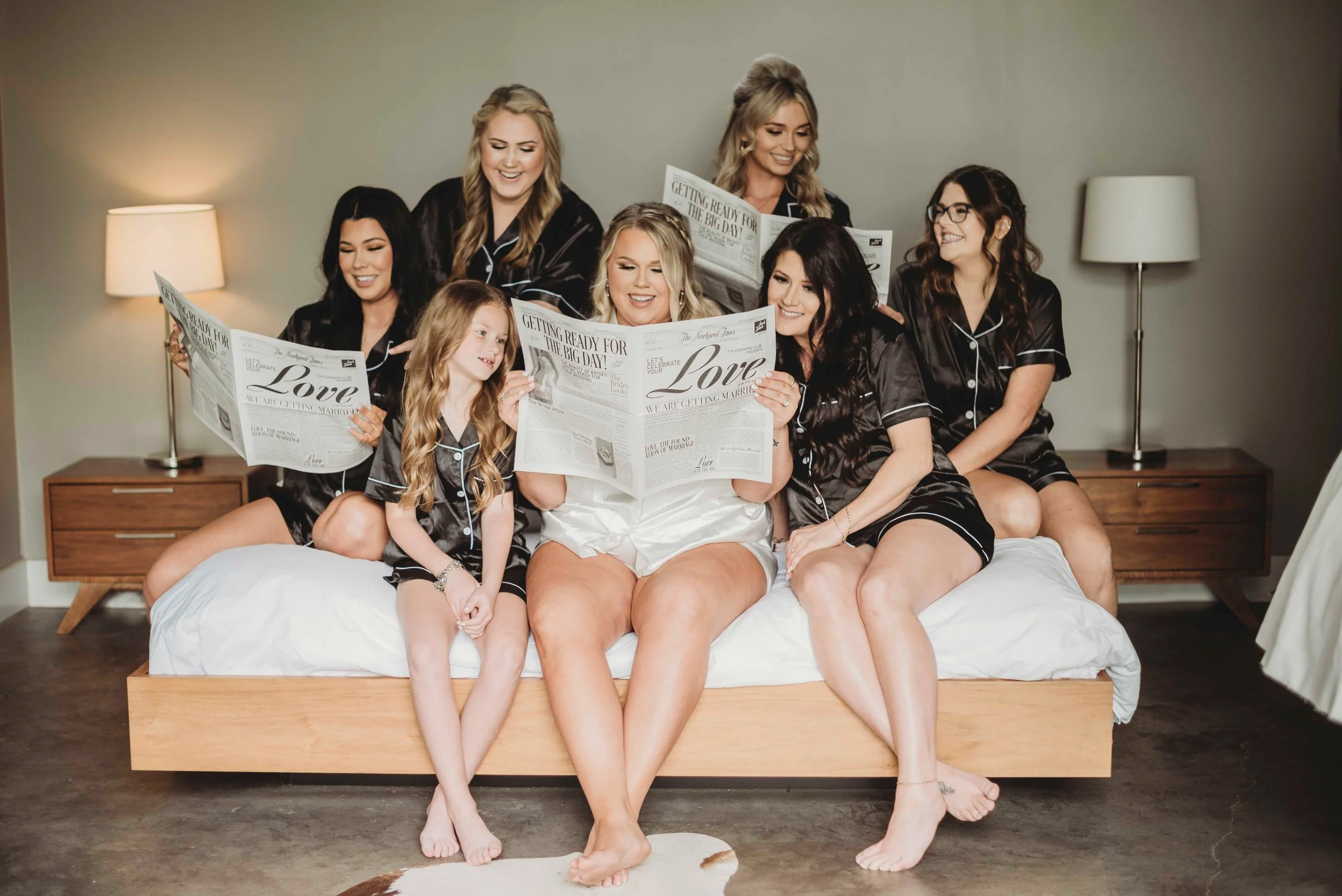 A group of women and a girl in matching pajamas sitting on a bed, reading newspapers, in a cozy bedroom setting with lamps on nightstands.