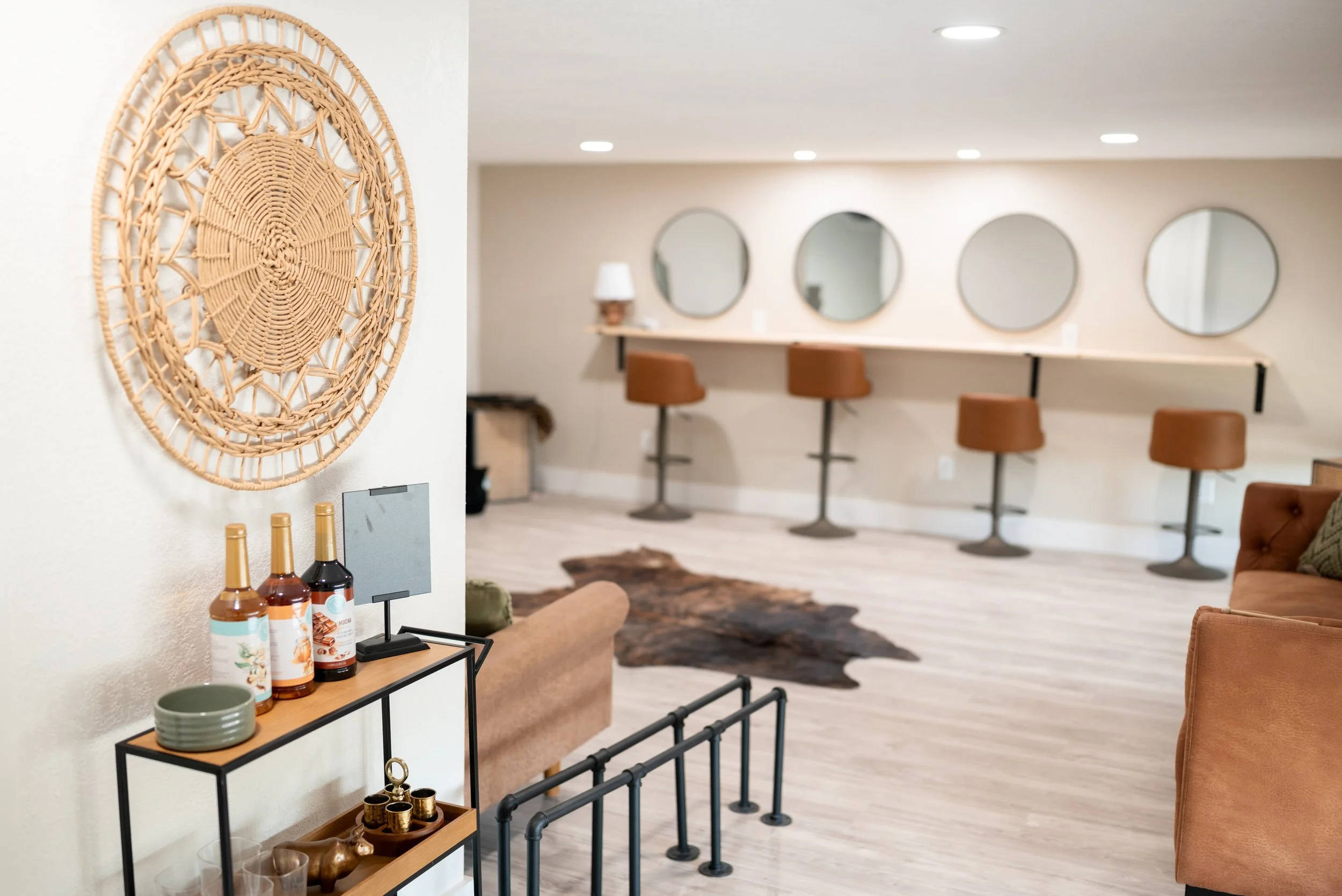 Interior of a lounge or bar area with four brown barstools at a counter with four mirrors on the wall, and a seating area with a sofa and decorative items including bottles and bowls.